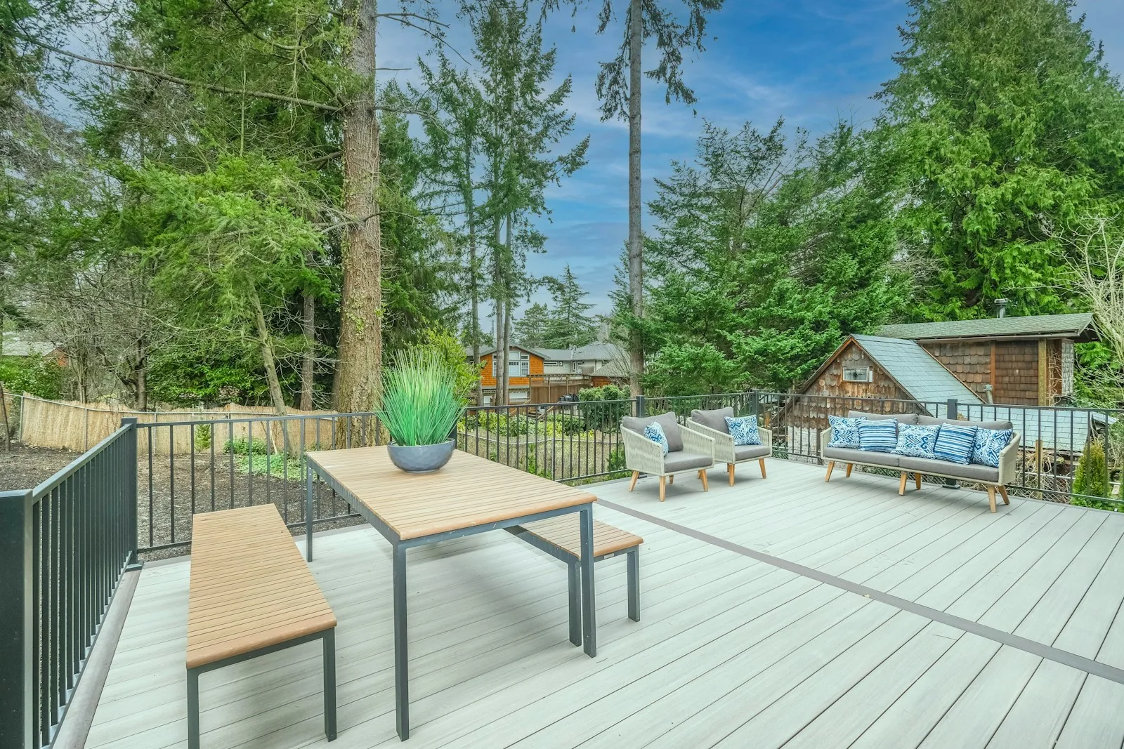 A spacious garden composite deck with grey decking boards, outdoor dining table, seating with cushions and metal railings, surrounded by green trees, representing the composite decking material option for above ground pool deck installations