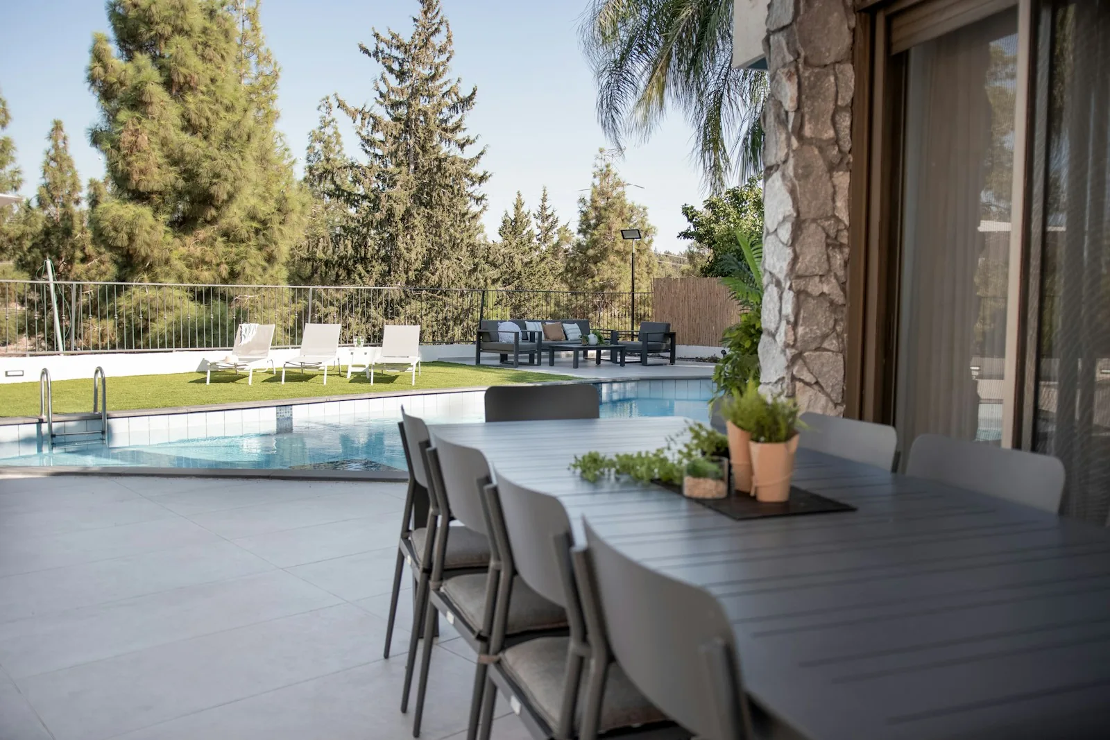 A modern outdoor entertaining space with grey dining table and chairs adjacent to a rectangular swimming pool with safety railings, representing the integrated outdoor living space created by adding decking or patio to a garden pool