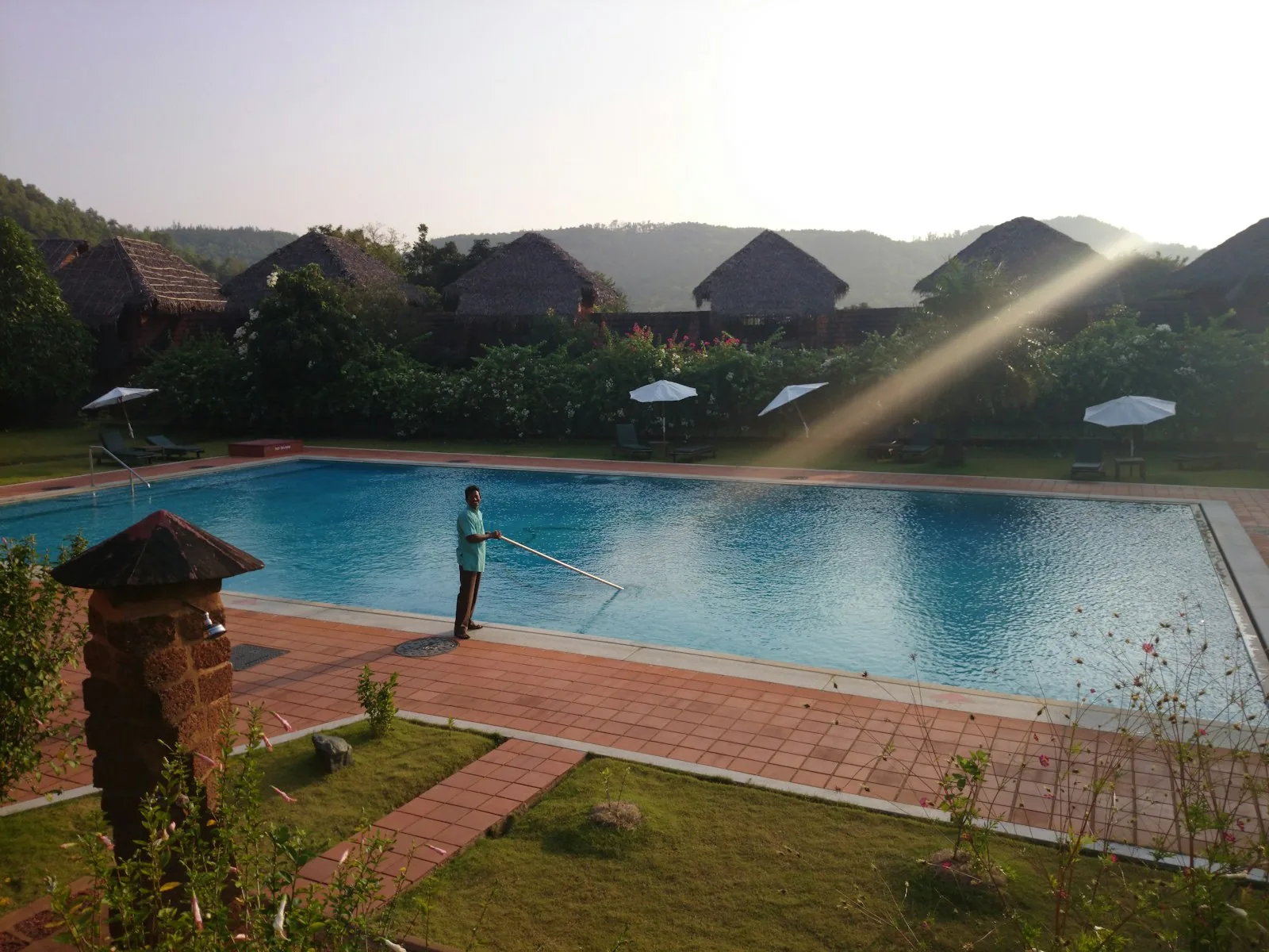 A pool maintenance worker cleaning a large outdoor swimming pool with a long-handled brush at sunrise, with tropical resort buildings in the background