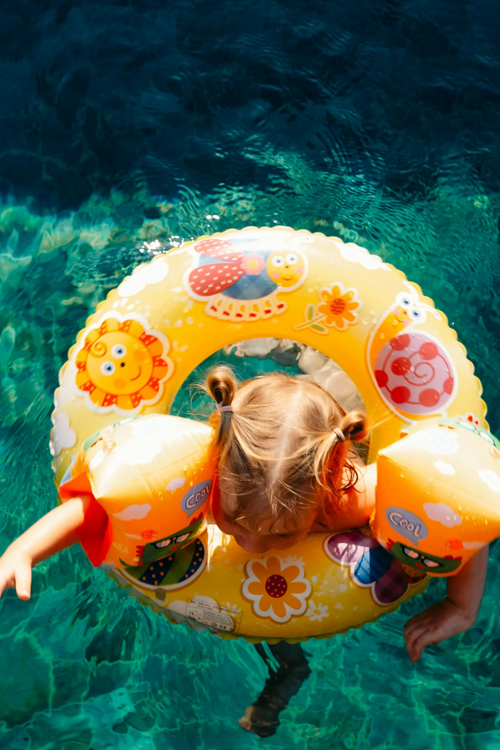 Overhead view of a young girl in a swimming pool wearing orange armbands and a yellow inflatable ring, representing the child safety considerations and supervision requirements for children using infinity pools and their catch basins