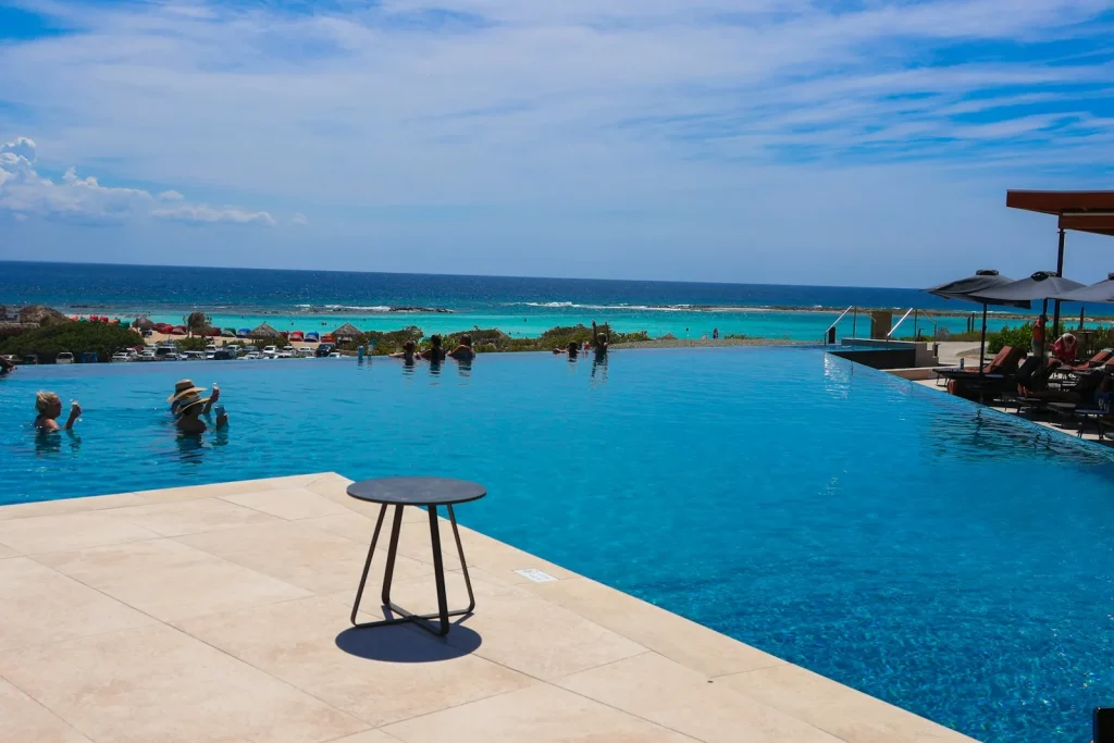 A resort infinity pool with people swimming in clear blue water, the vanishing edge merging visually with the turquoise Caribbean ocean beyond, showing the optical illusion created by the overflow weir wall design