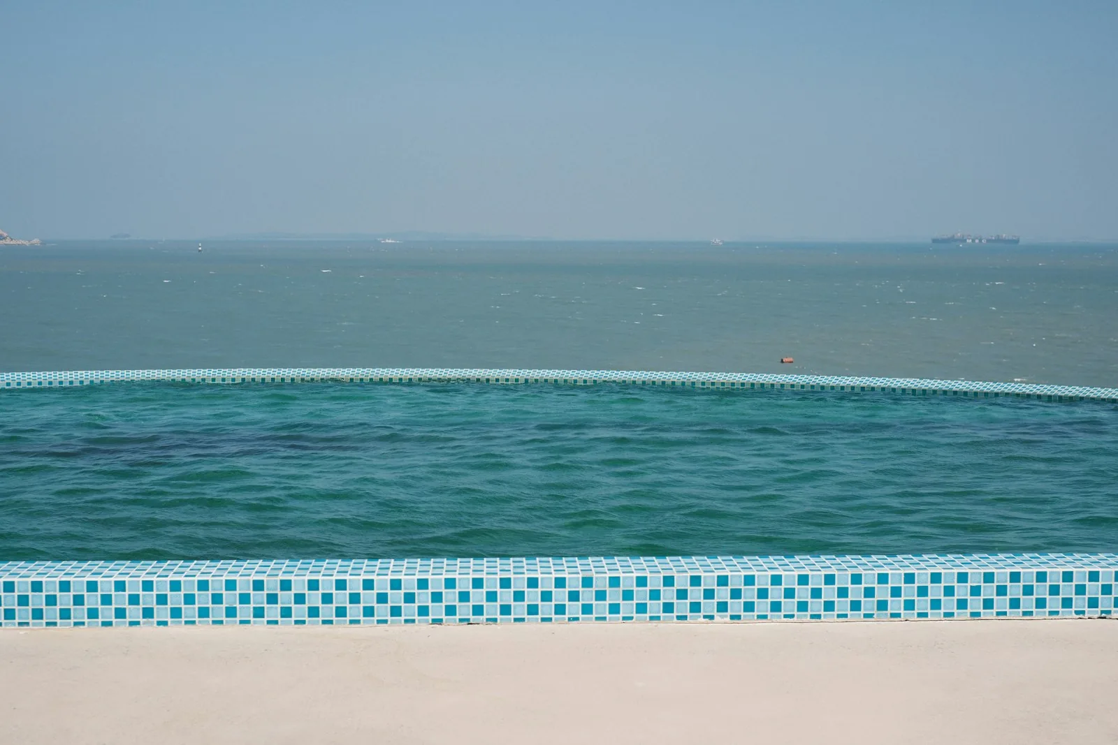 Close-up view of an infinity pool's vanishing overflow edge with blue and white mosaic tiles, water spilling over the weir wall toward the ocean beyond, illustrating the key safety design feature of infinity pool overflow systems