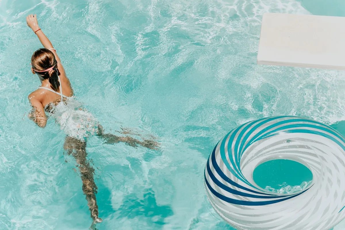 Overhead view of a girl swimming in a clear blue pool with a blue-and-white striped inflatable ring float, representing the family pool experience and practical information for visiting a Haven holiday park pool