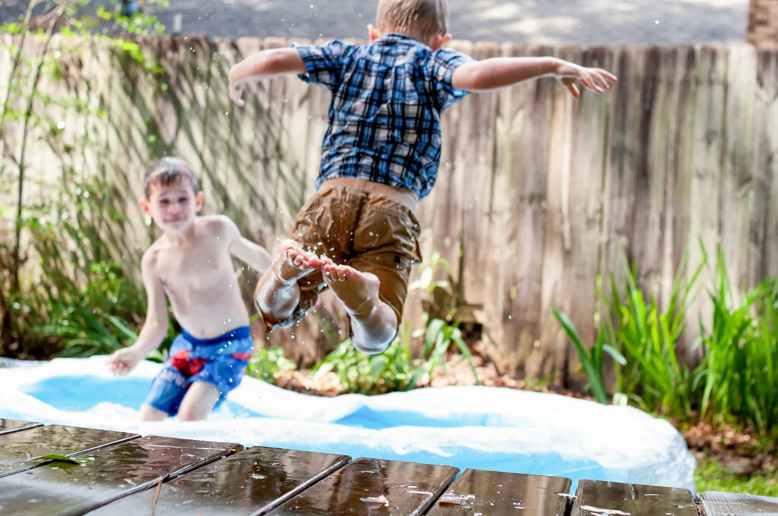 Two boys playing in an inflatable garden paddling pool in summer, one jumping in
