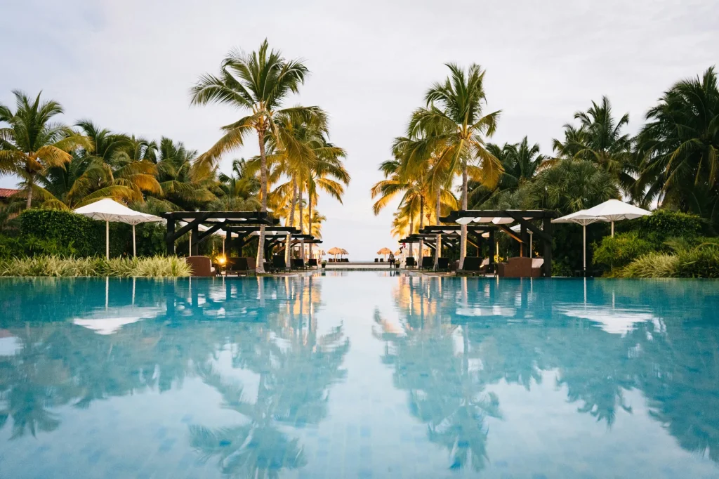 A large luxury resort pool at dusk surrounded by palm trees with white parasols and the water perfectly reflecting the scene