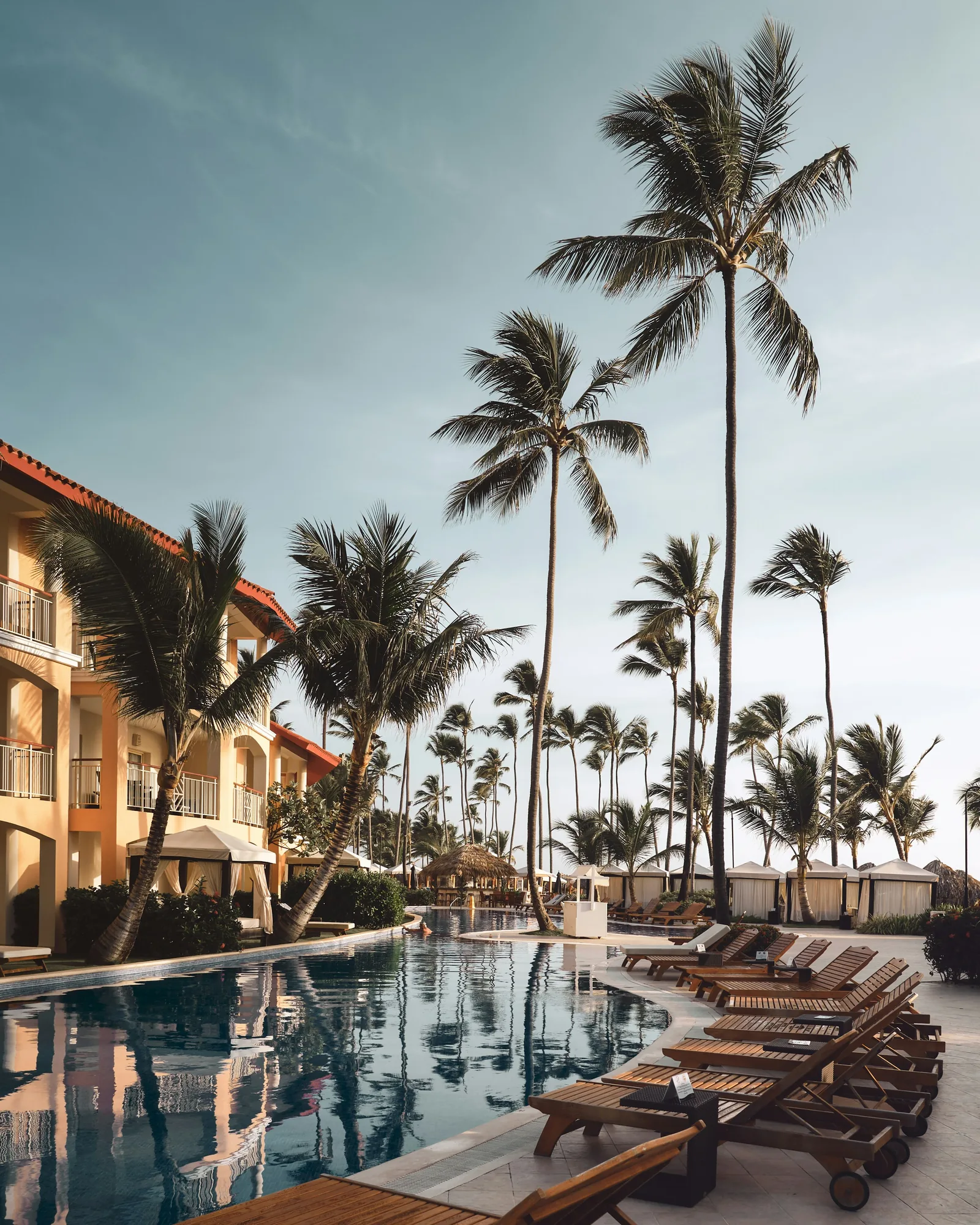 A resort hotel pool with tall palm trees swaying and wooden sun loungers alongside a yellow-painted hotel building