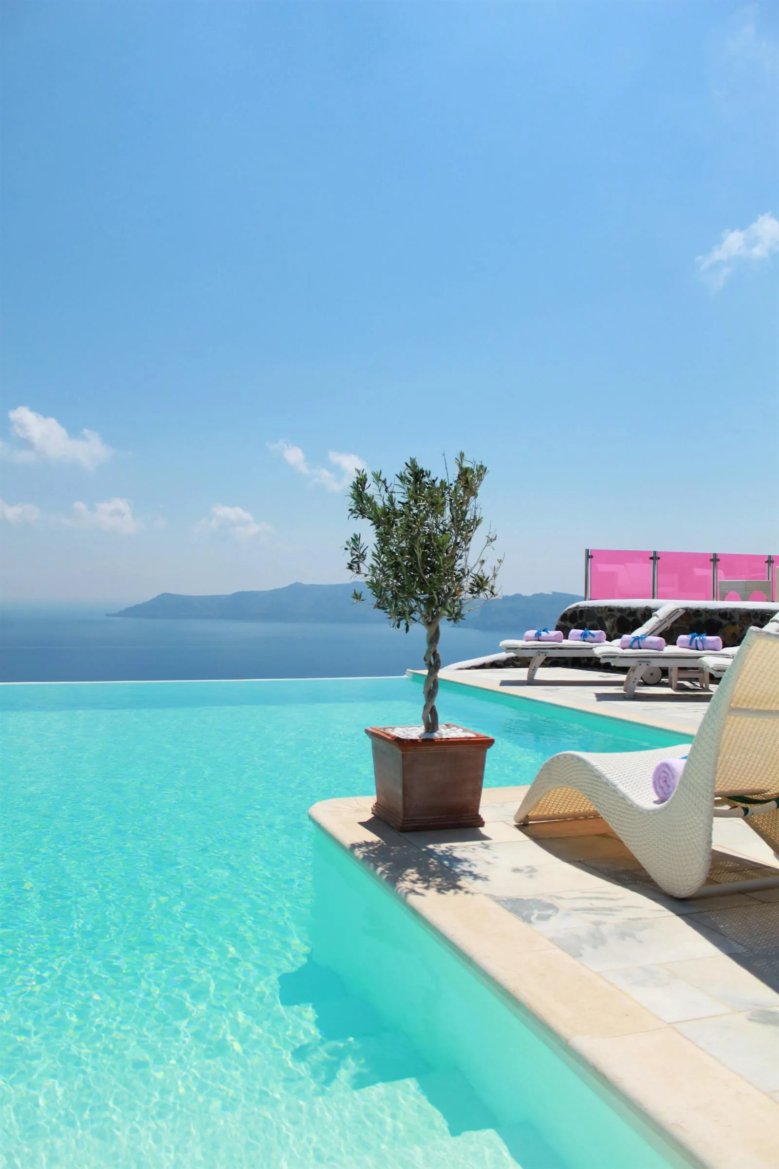 An infinity edge swimming pool at a cliffside hotel in Santorini Greece with the volcanic Caldera and Aegean Sea stretching to the horizon under a blue sky with white clouds, representing the caldera-view pool hotels in Imerovigli and Firostefani