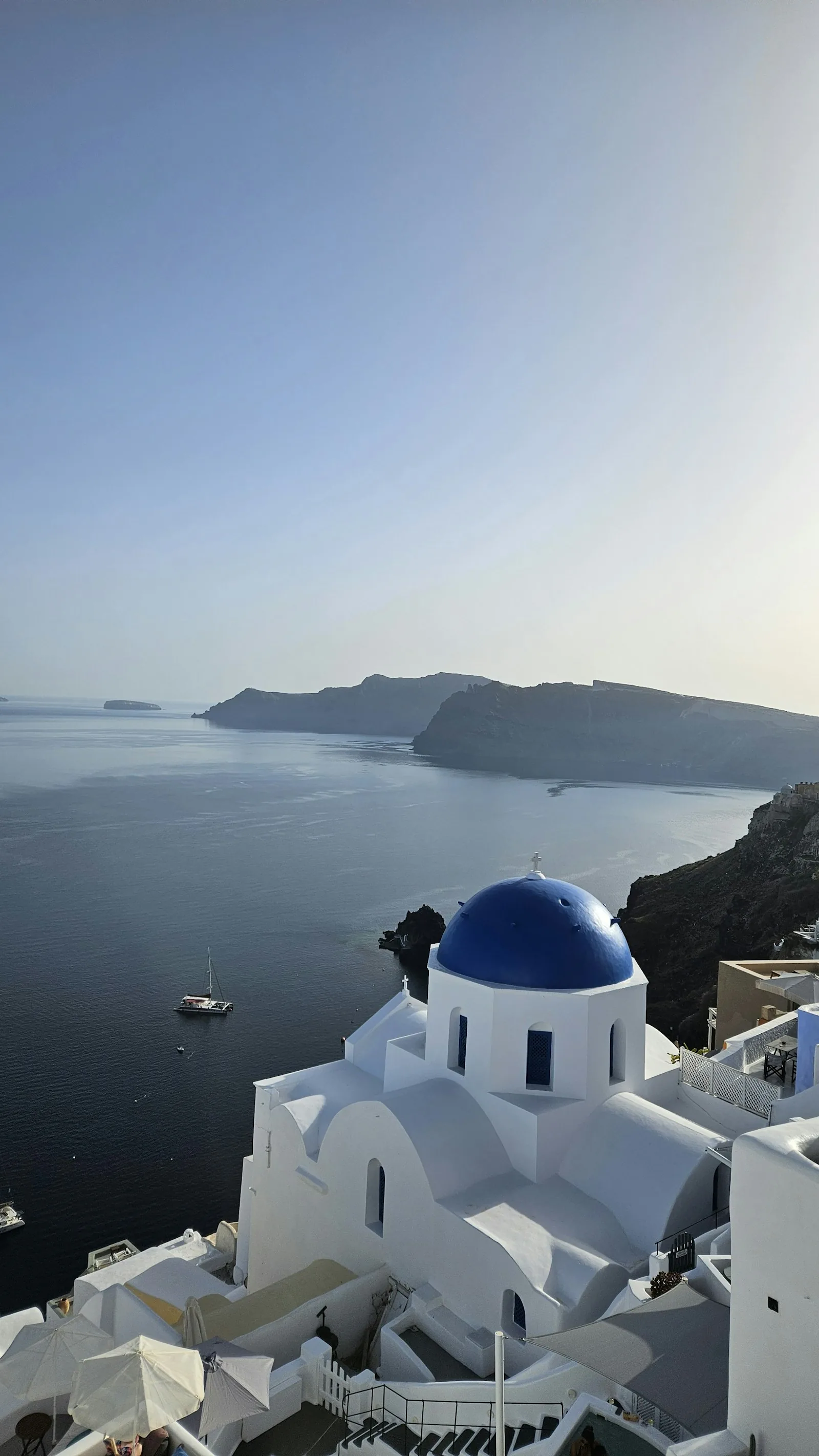 The white-washed architecture of Santorini Greece with blue-domed churches and terrace buildings cascading down a cliffside with views over the Aegean Sea, representing the Santorini village landscape and the location choices for hotel selection