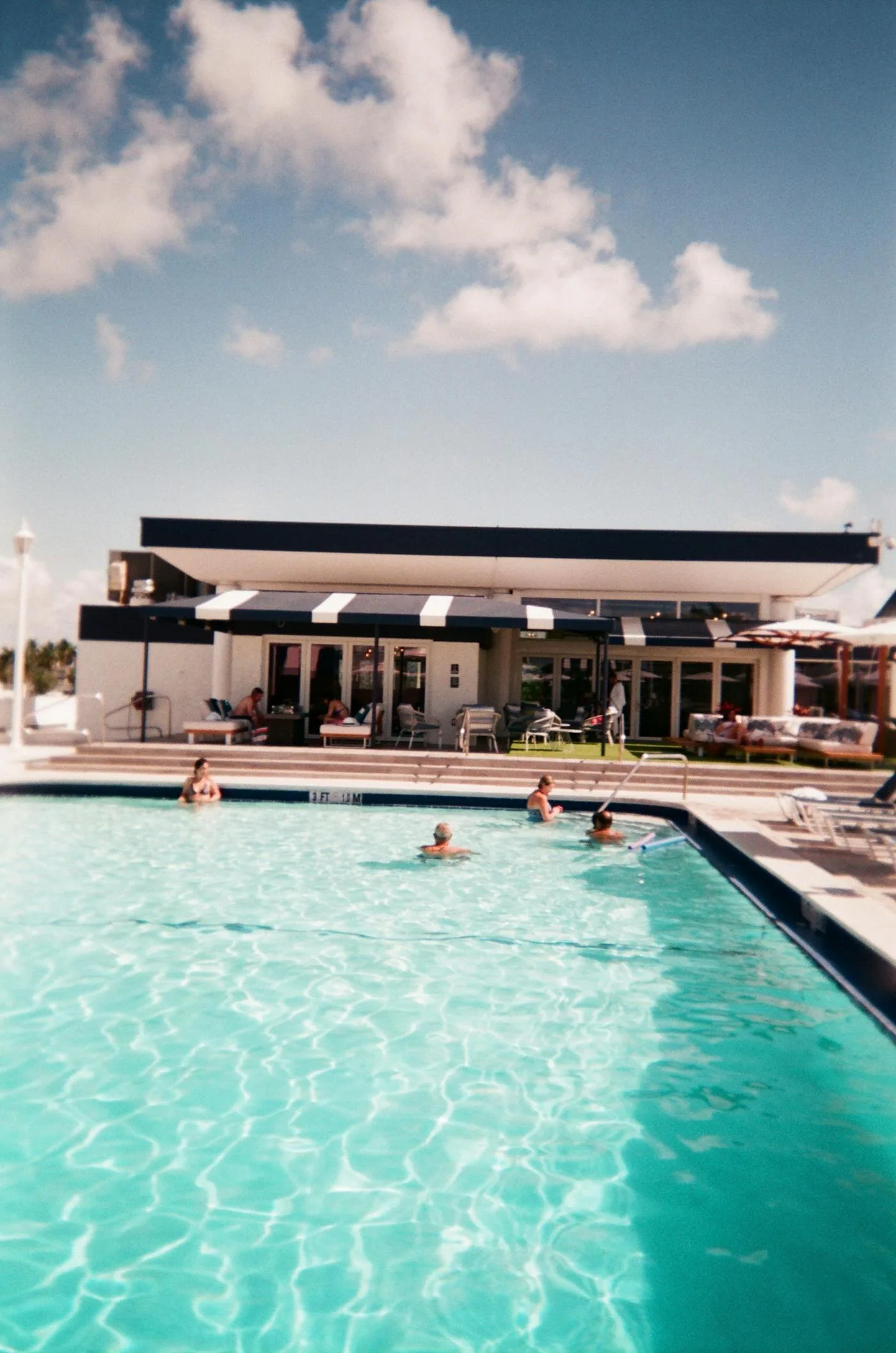 Guests swimming in an outdoor resort pool with sun loungers and a poolside building in the background