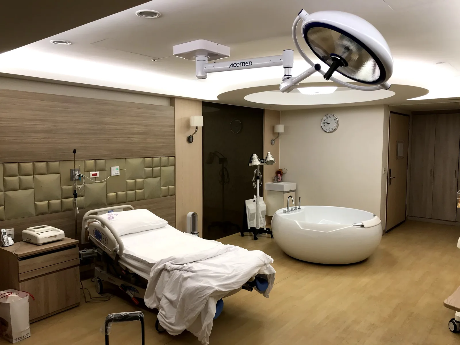 A modern hospital delivery room showing a round white birthing pool alongside a hospital bed and delivery lighting, representing water birth facilities