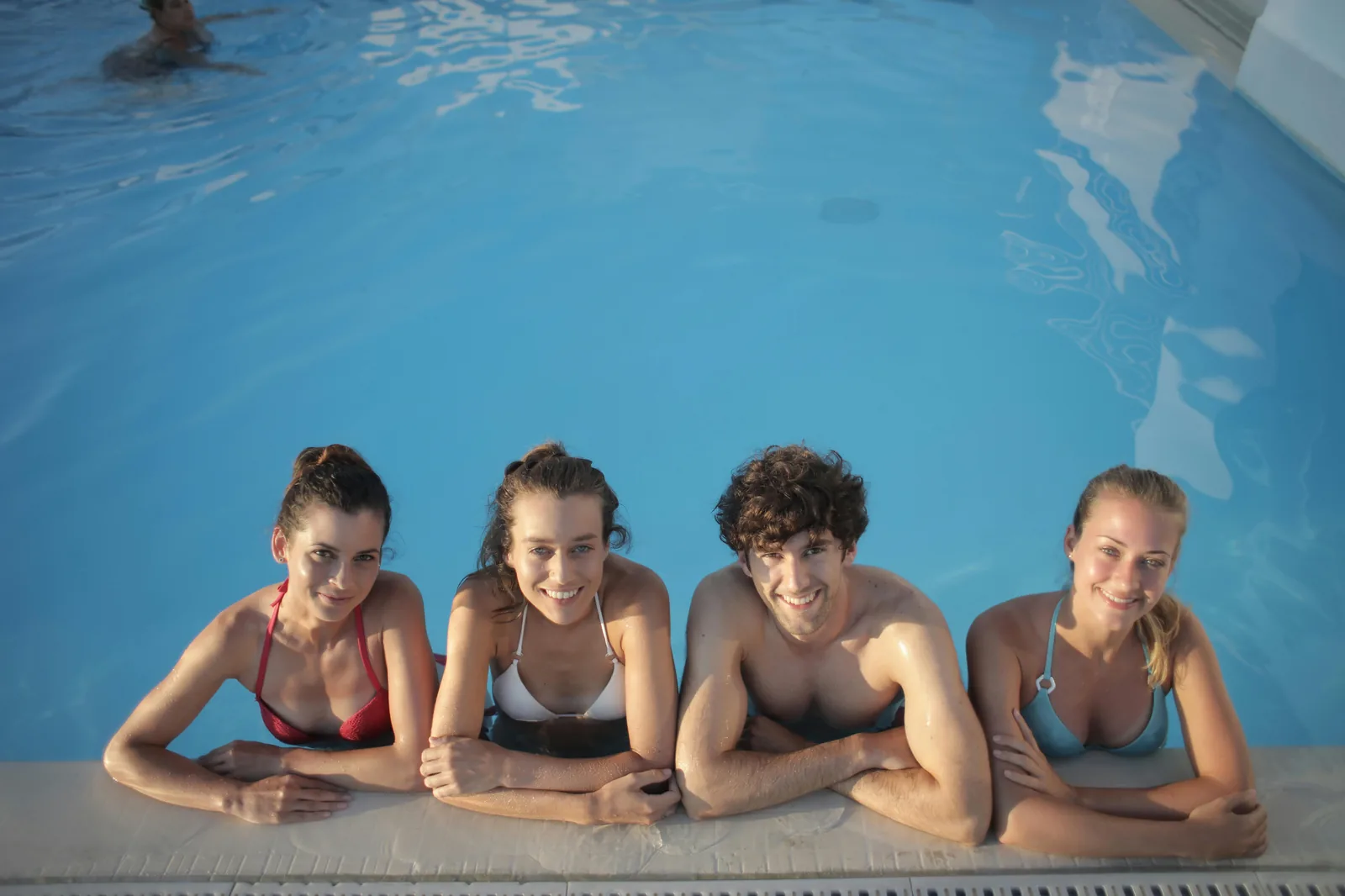 Four smiling friends leaning on the edge of a bright blue swimming pool