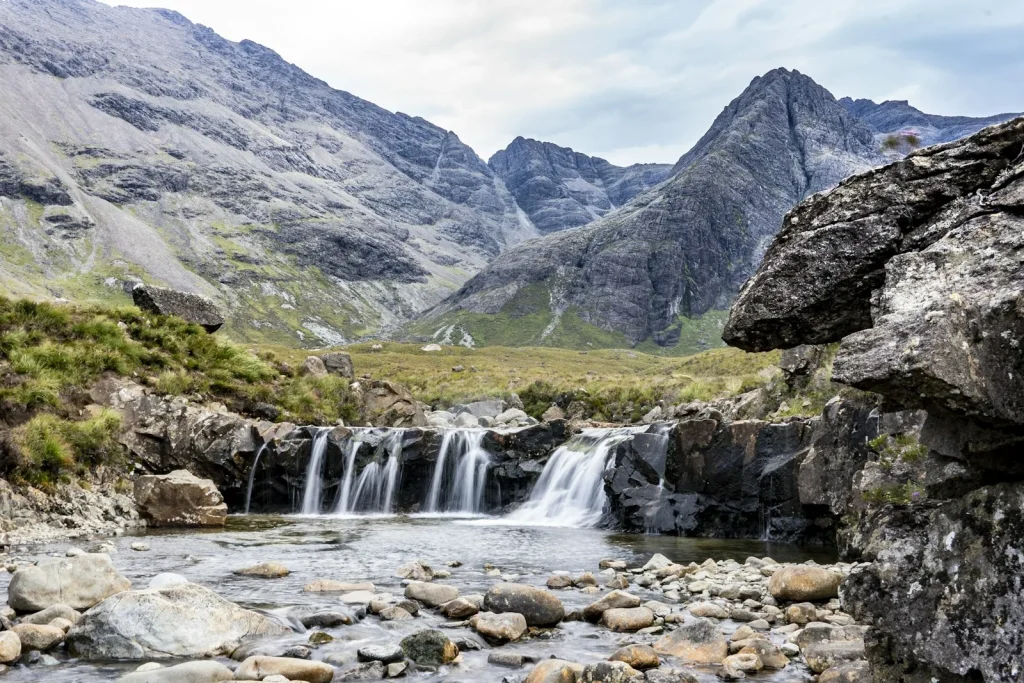 A natural waterfall cascading over rocks into a clear pool in a Scottish highland landscape with dramatic Black Cuillin mountains behind, representing the type of fairy pools and natural wild swimming spots found in Scotland including near Luss on Loch Lomond