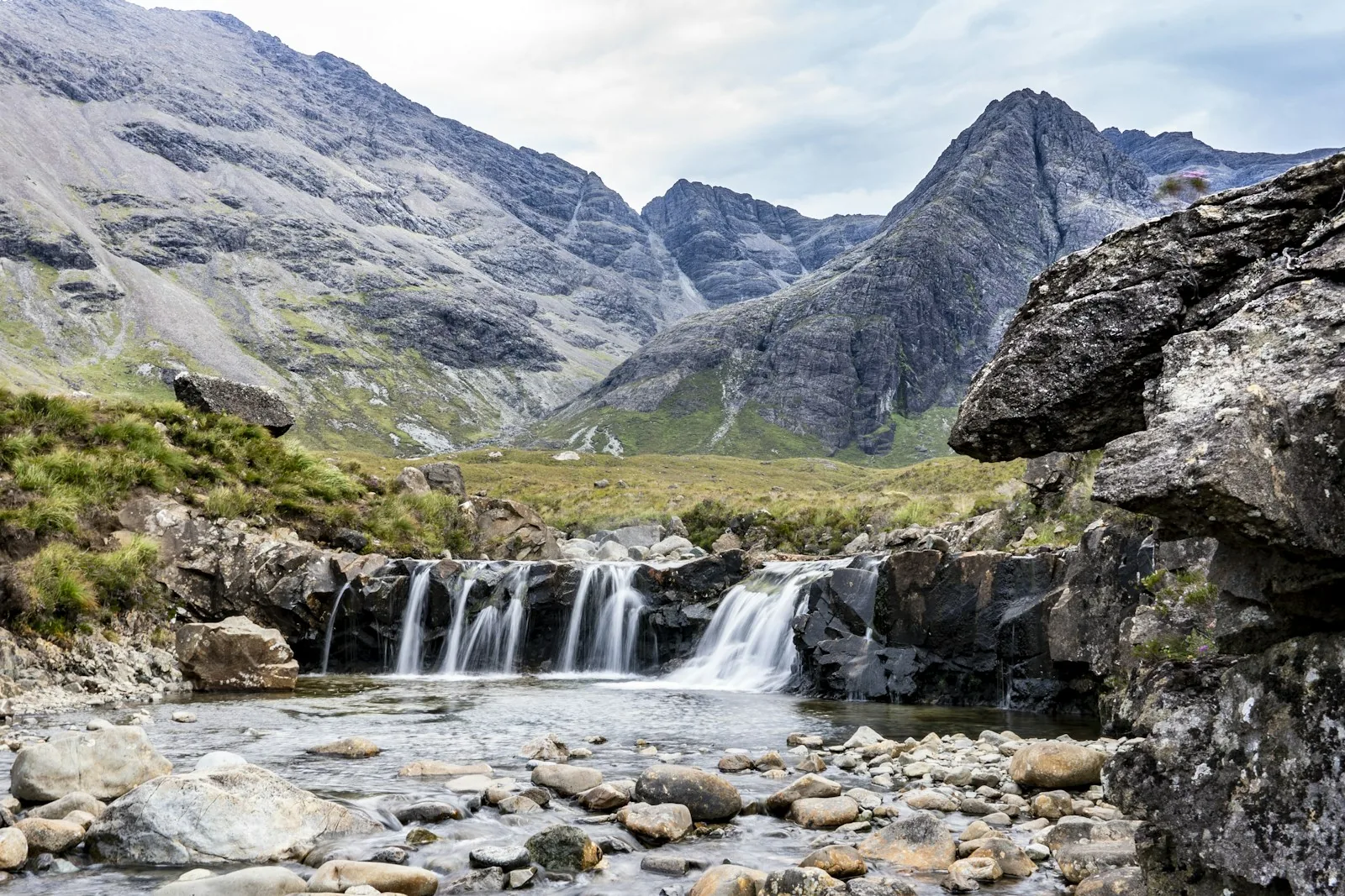 A natural waterfall cascading over rocks into a clear pool in a Scottish highland landscape with dramatic Black Cuillin mountains behind, representing the type of fairy pools and natural wild swimming spots found in Scotland including near Luss on Loch Lomond