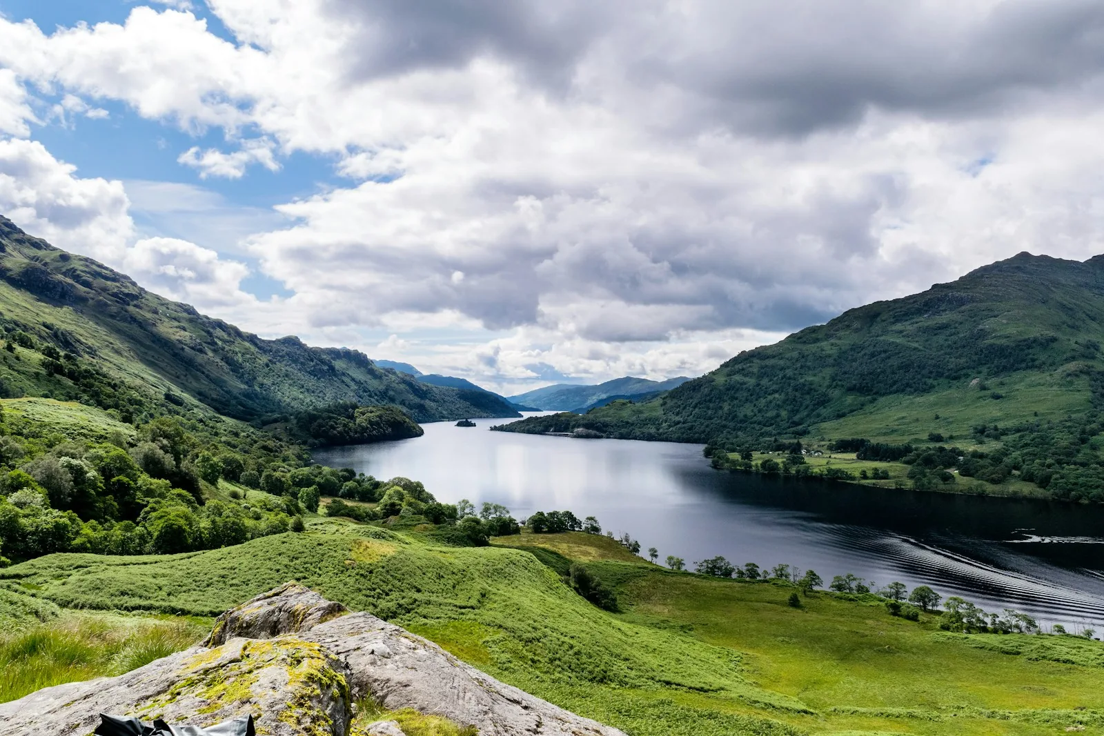 Elevated panoramic view of Loch Lomond from a hillside viewpoint showing the full length of the loch stretching between green hills and mountains under a partly cloudy sky, representing the landscape of the Luss and Loch Lomond area for visitors planning a trip