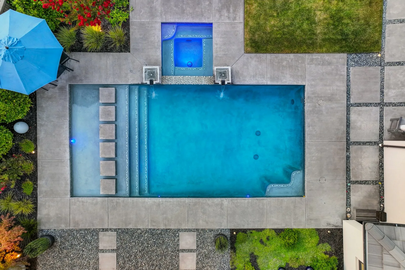 An aerial drone view of a rectangular inground pool with integrated spa, stepping stones and landscaped garden surround