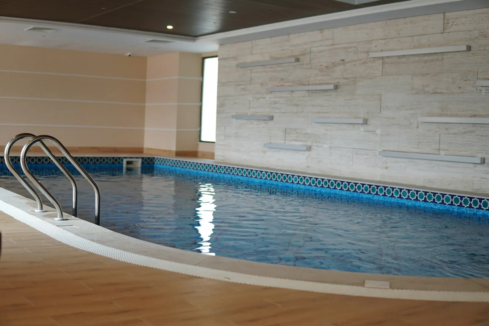 An empty indoor health club swimming pool with clear blue water, decorative mosaic tile border, stone wall cladding, and pool entry steps, representing the indoor pool facilities at Edinburgh leisure centres and private health clubs