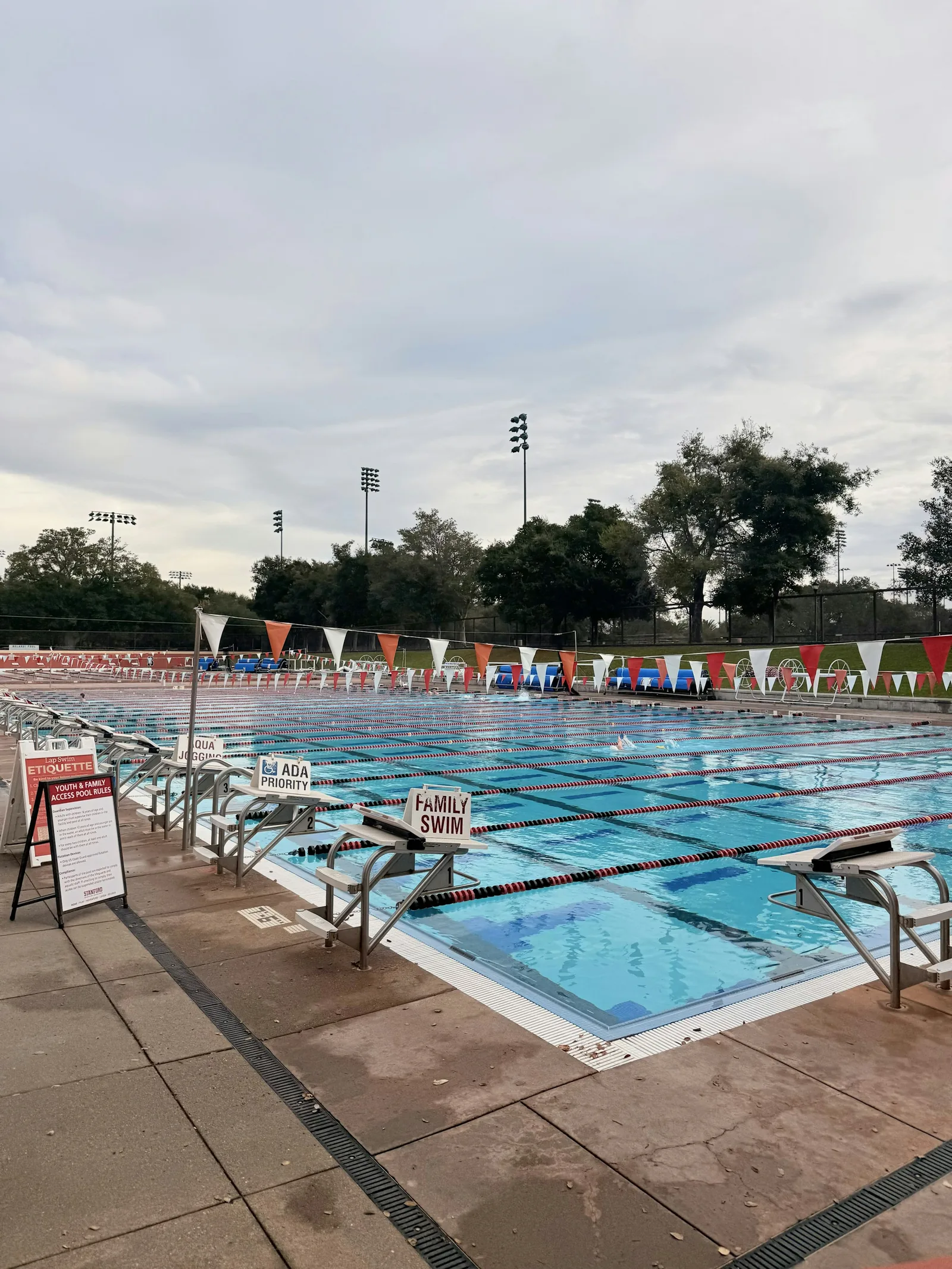 An outdoor multi-lane competition swimming pool with diving blocks, lane markers, family swim signs, and overcast sky, representing the outdoor pool facilities available at premium Edinburgh gym memberships including David Lloyd Edinburgh Shawfair