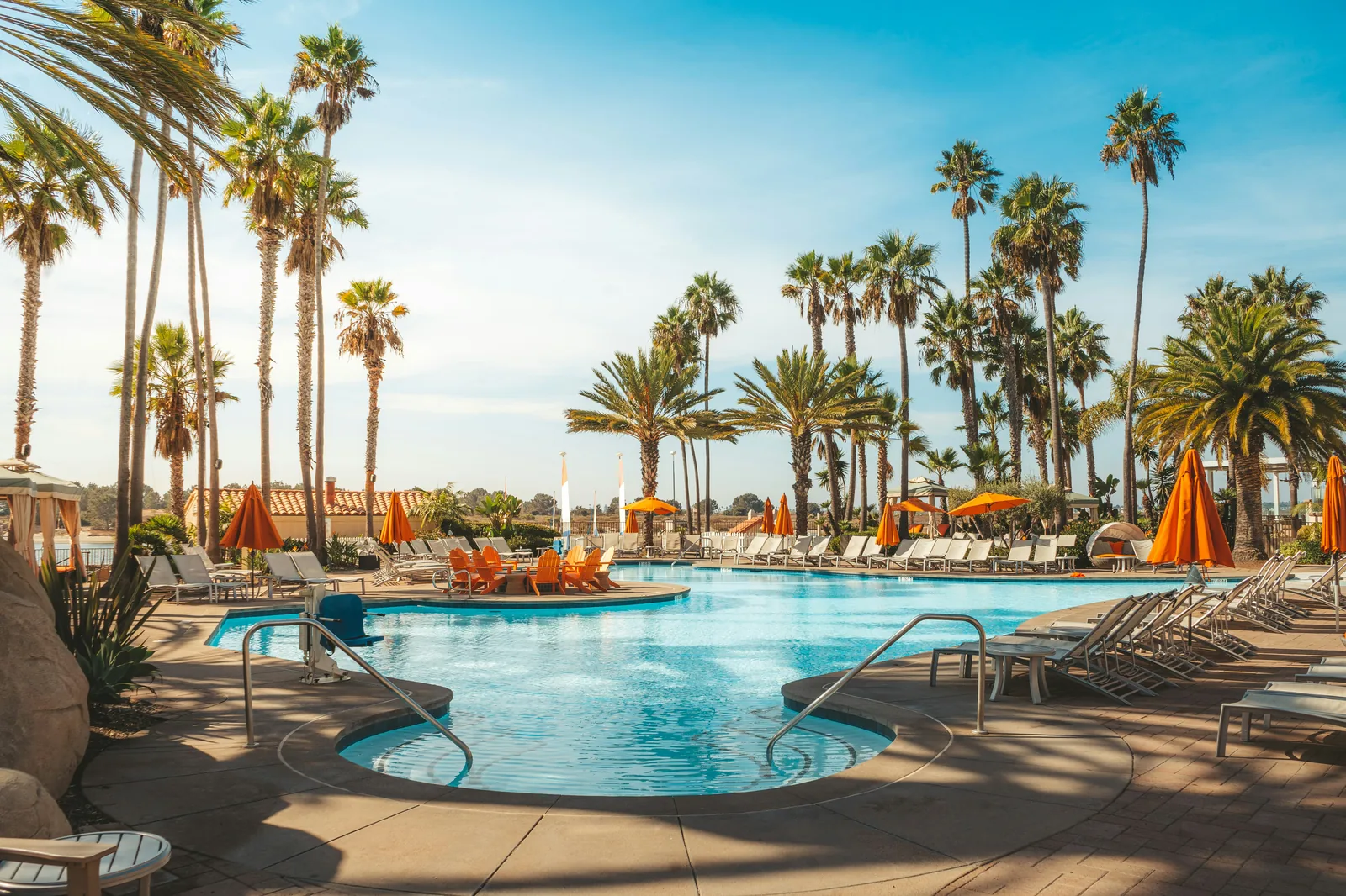 A large outdoor swimming pool at a resort with orange umbrellas, sun loungers and tall palm trees