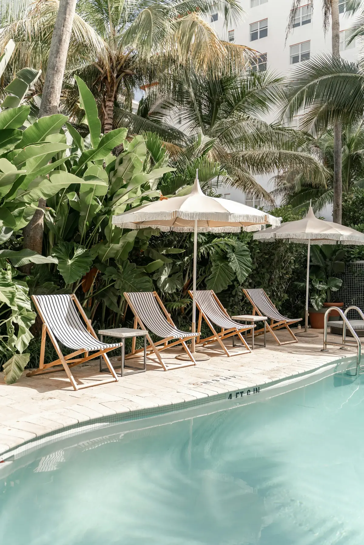 An outdoor hotel pool with striped wooden loungers, fringed parasols and lush tropical greenery