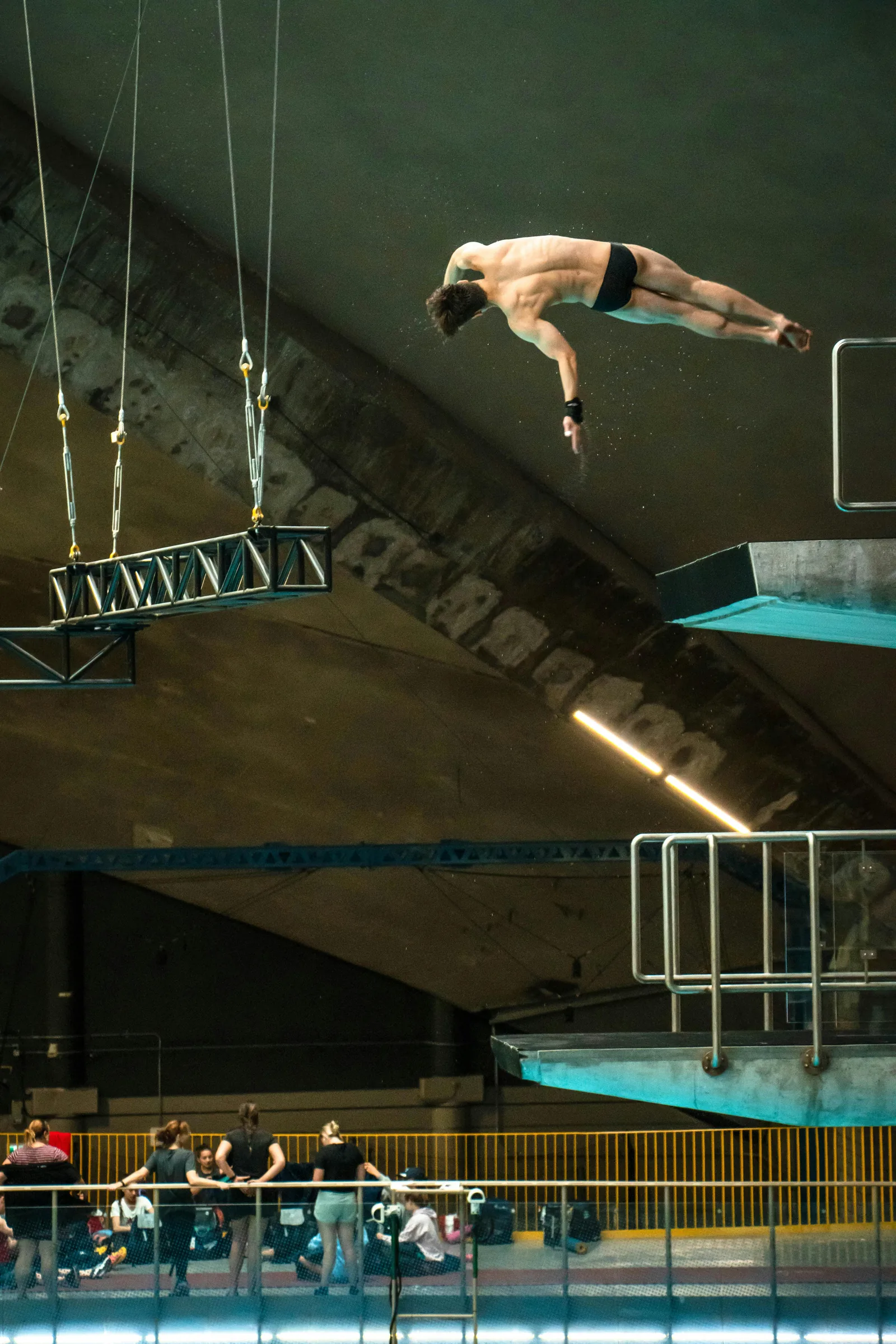 A competitive diver launching off a high platform diving board at an Olympic aquatics centre above a deep competition diving pool representing the 5 metre minimum depth requirement for 10 metre platform diving pools