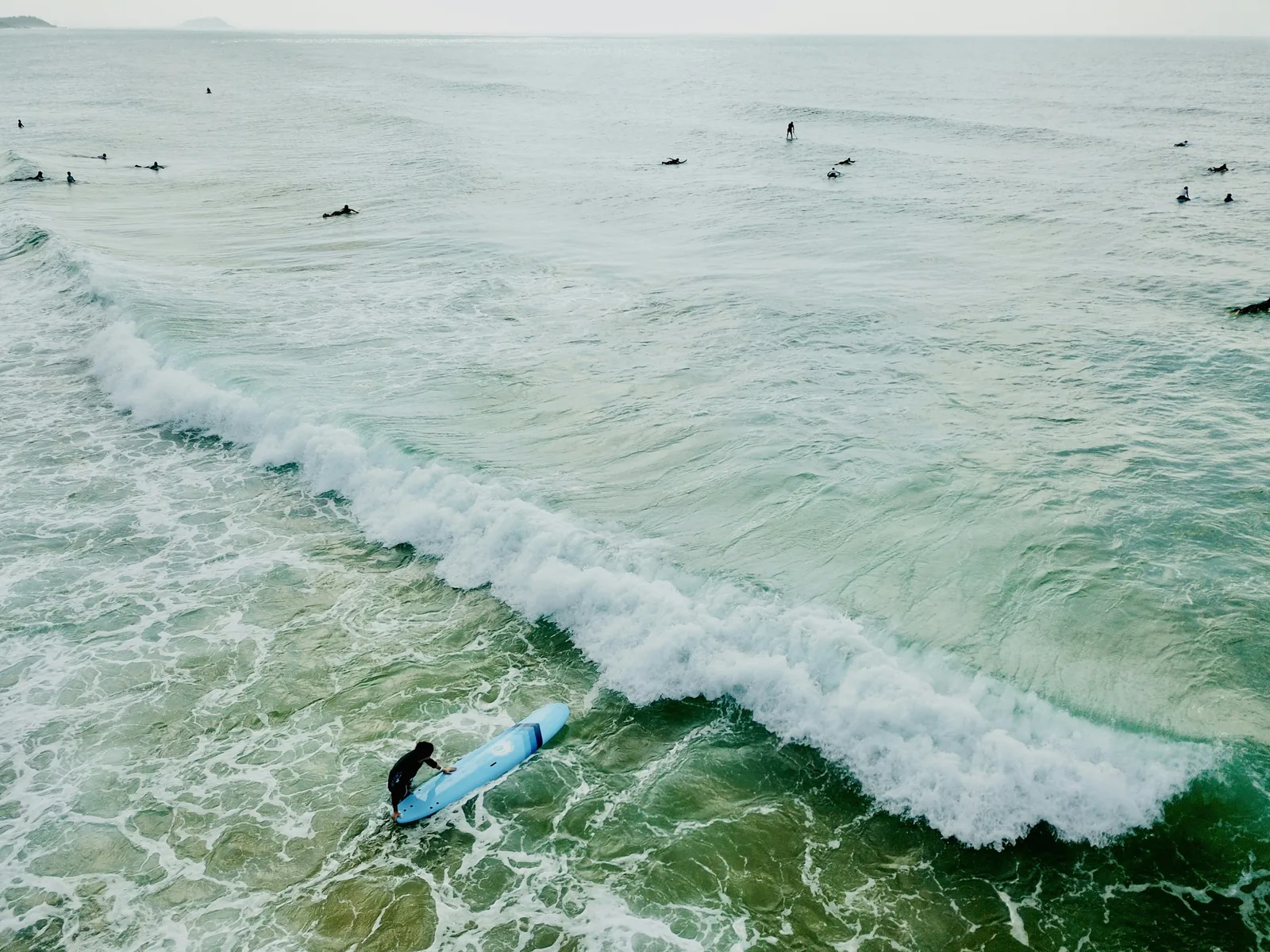 Aerial drone view of a breaking wave with multiple surfers and bodyboarders in the water, illustrating how pool bathymetry and wave shape determine the character and size of surf waves