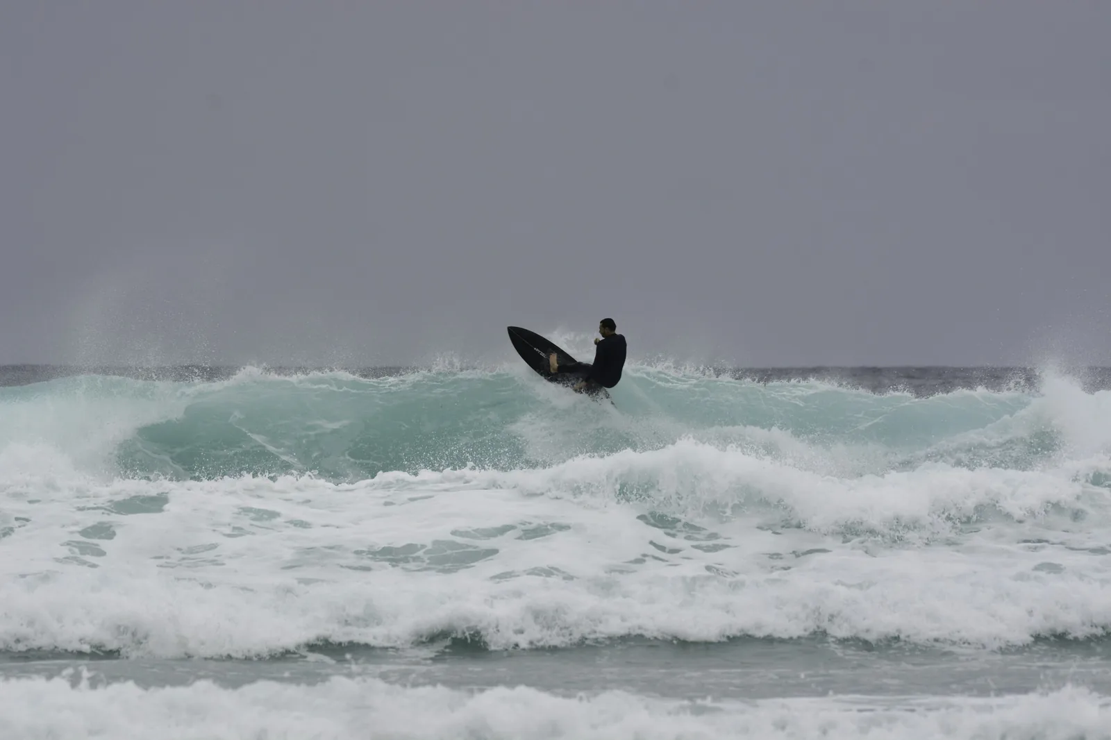 A surfer riding a breaking wave at Watergate Bay Newquay UK in grey overcast conditions, representing UK surfing and the wave pool facilities available at The Wave Bristol and Lost Shore Edinburgh