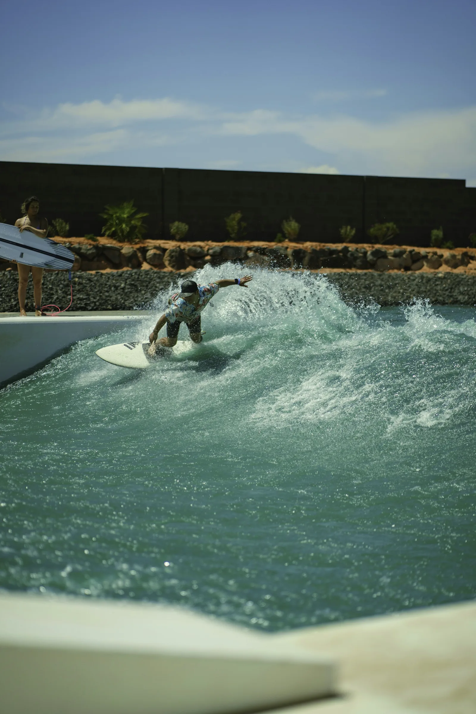 A surfer riding an artificial wave at a wave pool facility with concrete perimeter walls and desert landscape behind, representing the pneumatic and mechanical wave generation technologies used in surf parks