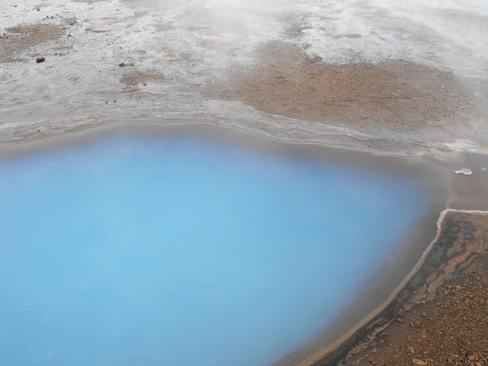 Milky turquoise-blue geothermal pool in the Geysir area of Iceland surrounded by mineral-encrusted volcanic rock, representing the characteristic silica-rich water found in Iceland's major commercial geothermal lagoons including the Blue Lagoon