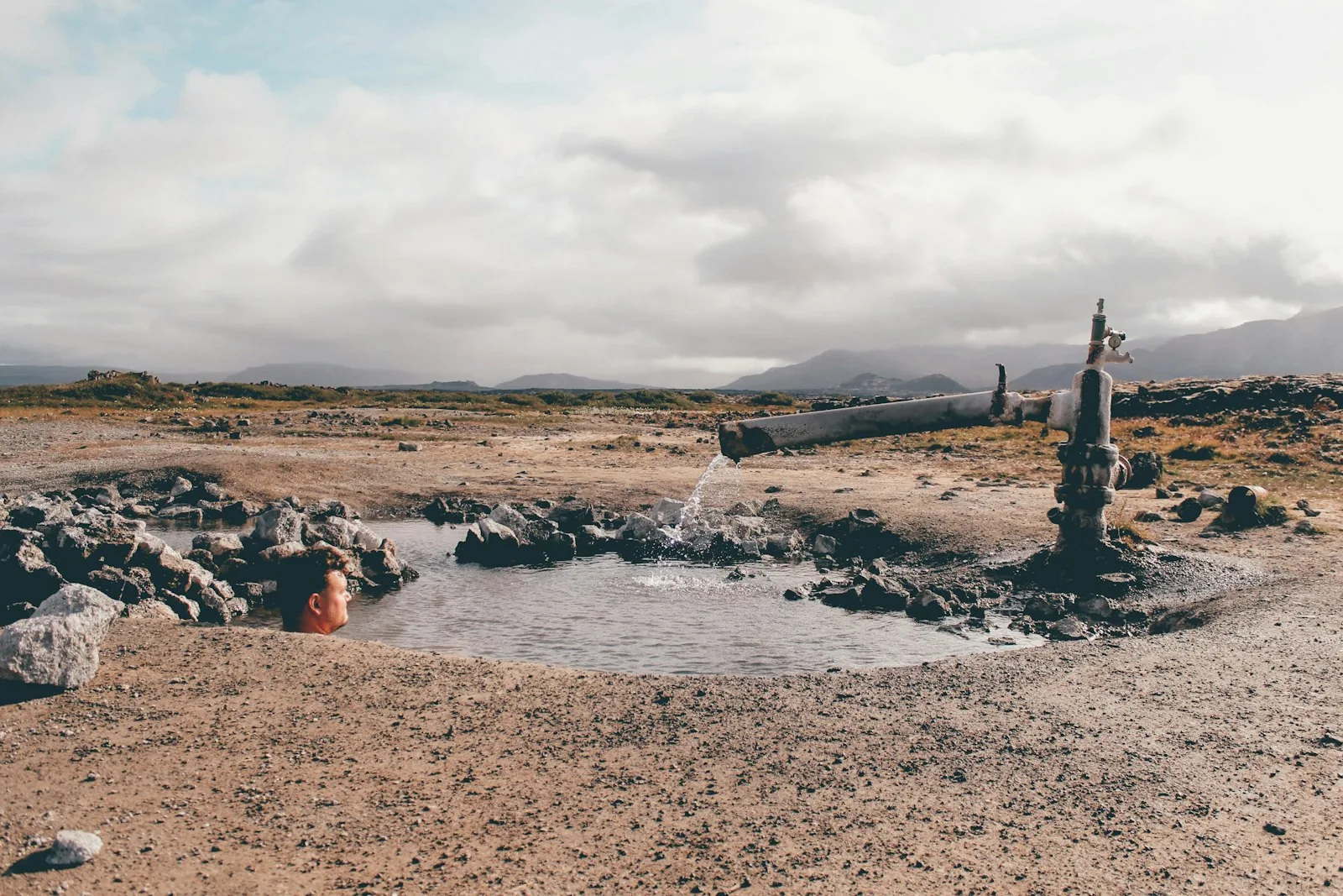 A person immersed in a small natural geothermal pool fed by a pipe on Iceland's volcanic terrain, representing the country's deep-rooted culture of outdoor geothermal bathing in sundlaugar and the accessible hot springs found across the island