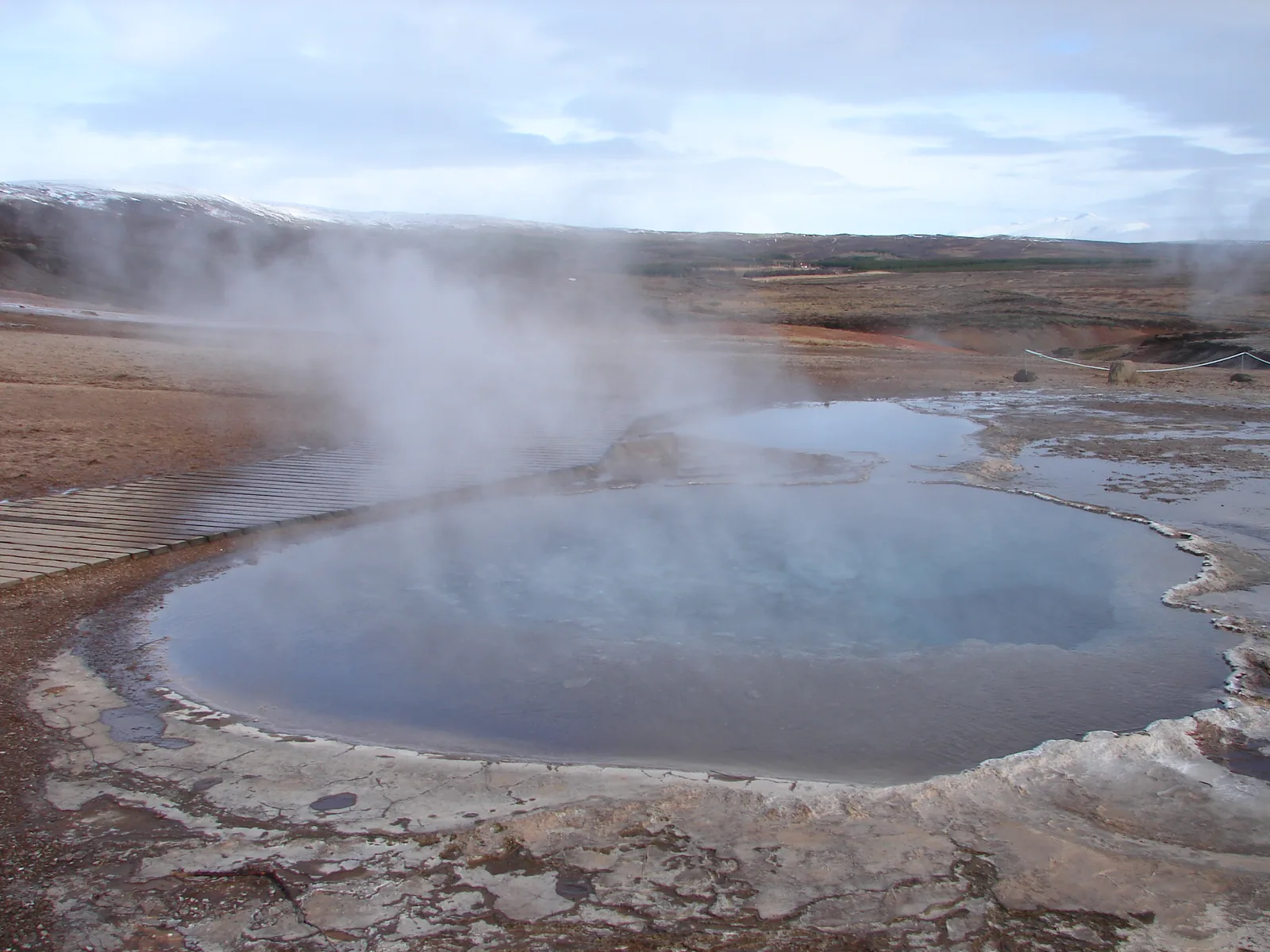 A steaming geothermal pool surrounded by mineral-encrusted volcanic rock with steam rising above Iceland's open moorland, representing the natural hot springs and wild geothermal bathing accessible to visitors away from commercial developments