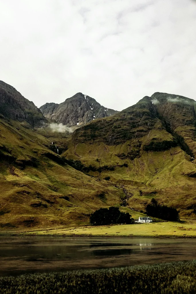A lone white cottage in a Scottish Highland glen with dramatic mountains and a loch in the foreground