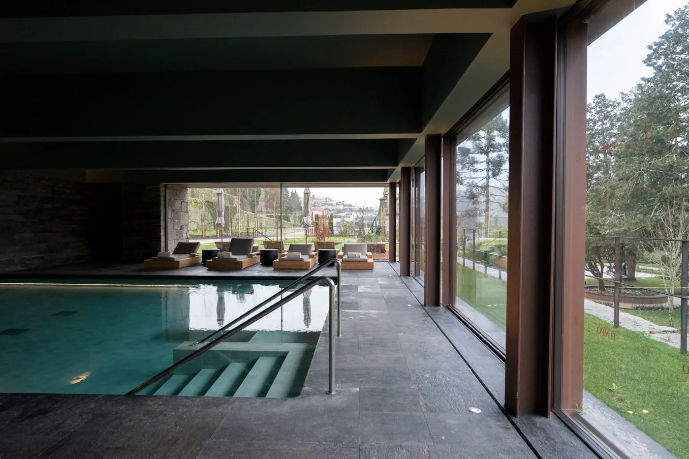 A luxury indoor swimming pool with floor-to-ceiling glass walls overlooking a garden, with lounge chairs alongside