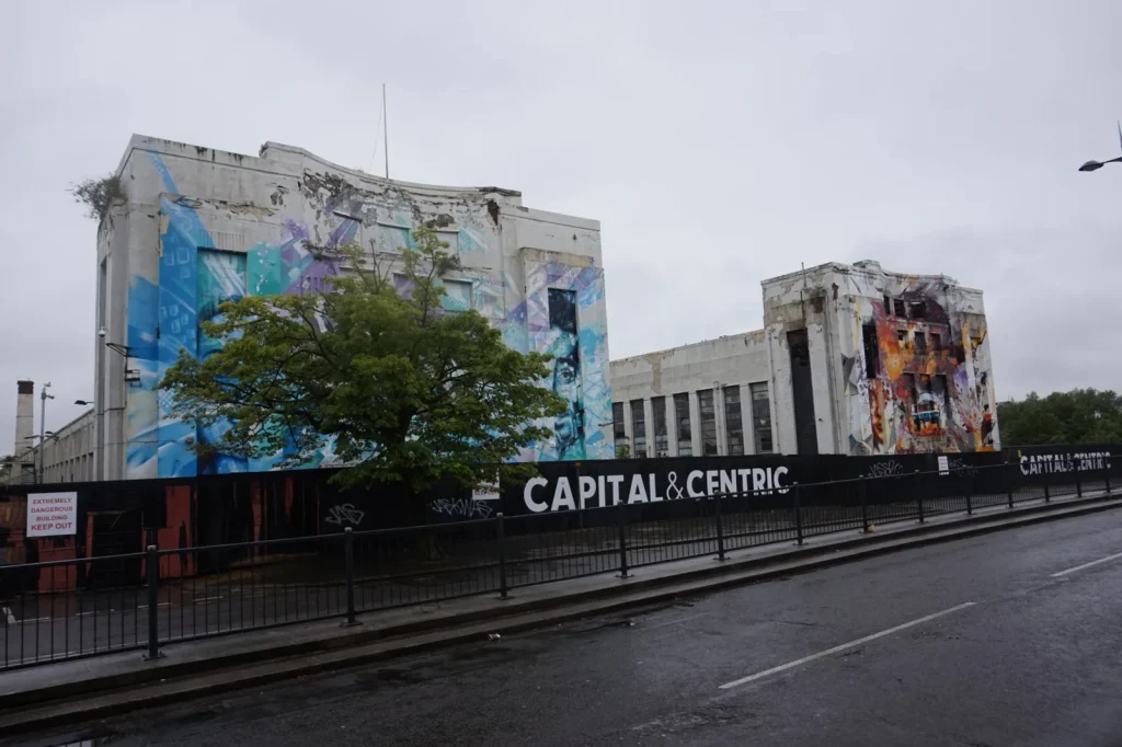 The Littlewoods Pools building on Edge Lane in Liverpool showing the white Art Deco stucco facade decorated with large painted murals, with Capital and Centric construction hoardings at street level and a danger sign, representing the building's pre-redevelopment state