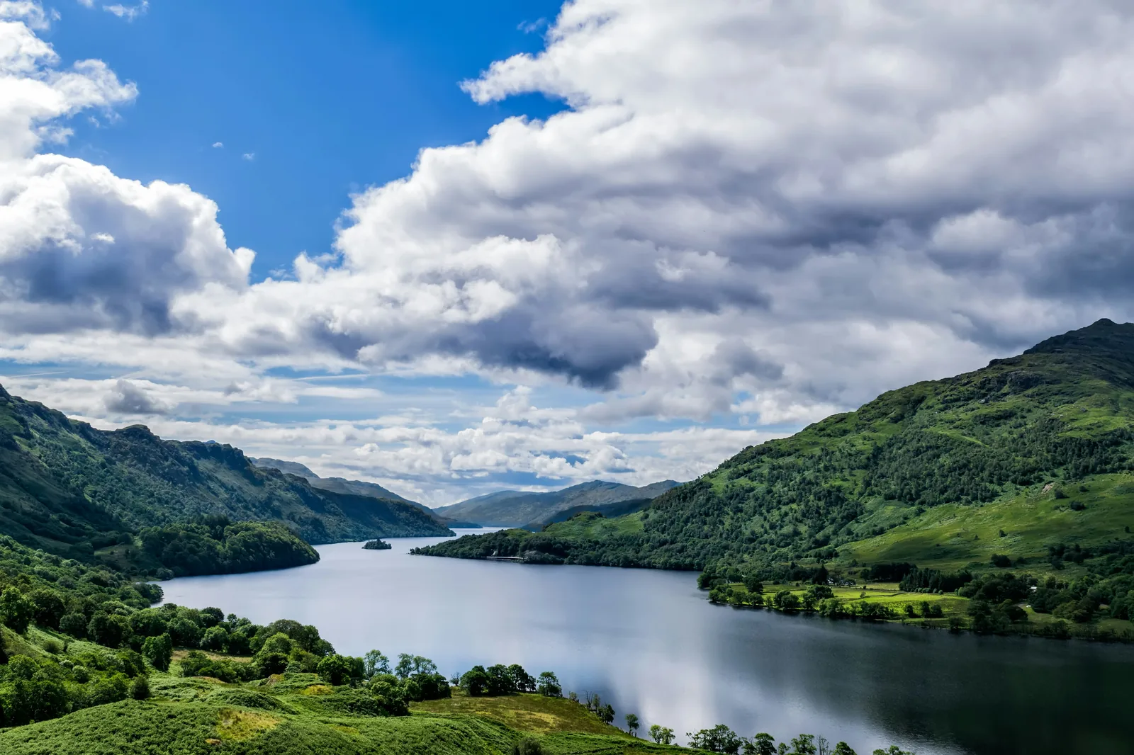 Aerial view of a Scottish loch flanked by green mountains under a dramatic blue sky with white clouds