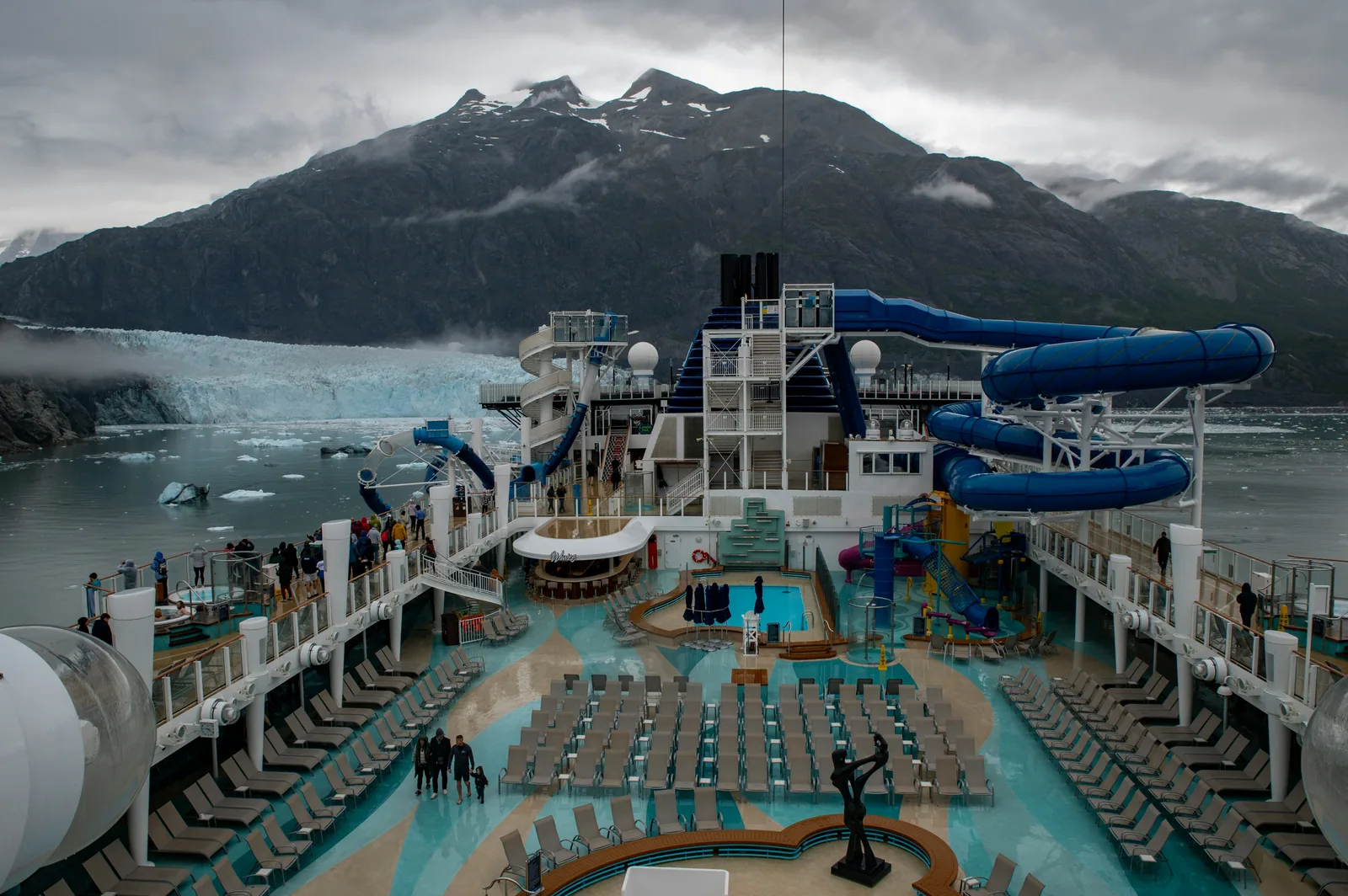 Cruise ship aquapark showing large blue water slides and pool deck with mountain backdrop