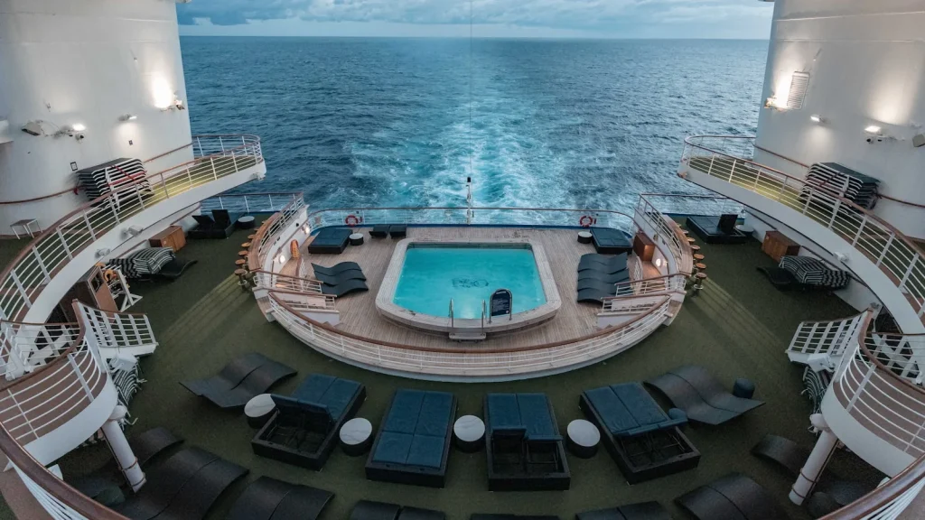 Aerial view of a cruise ship outdoor pool on the aft deck surrounded by dark sunloungers on a teak deck, with the open sea and wake visible behind the ship, representing the outdoor swimming pool experience on P&O Britannia