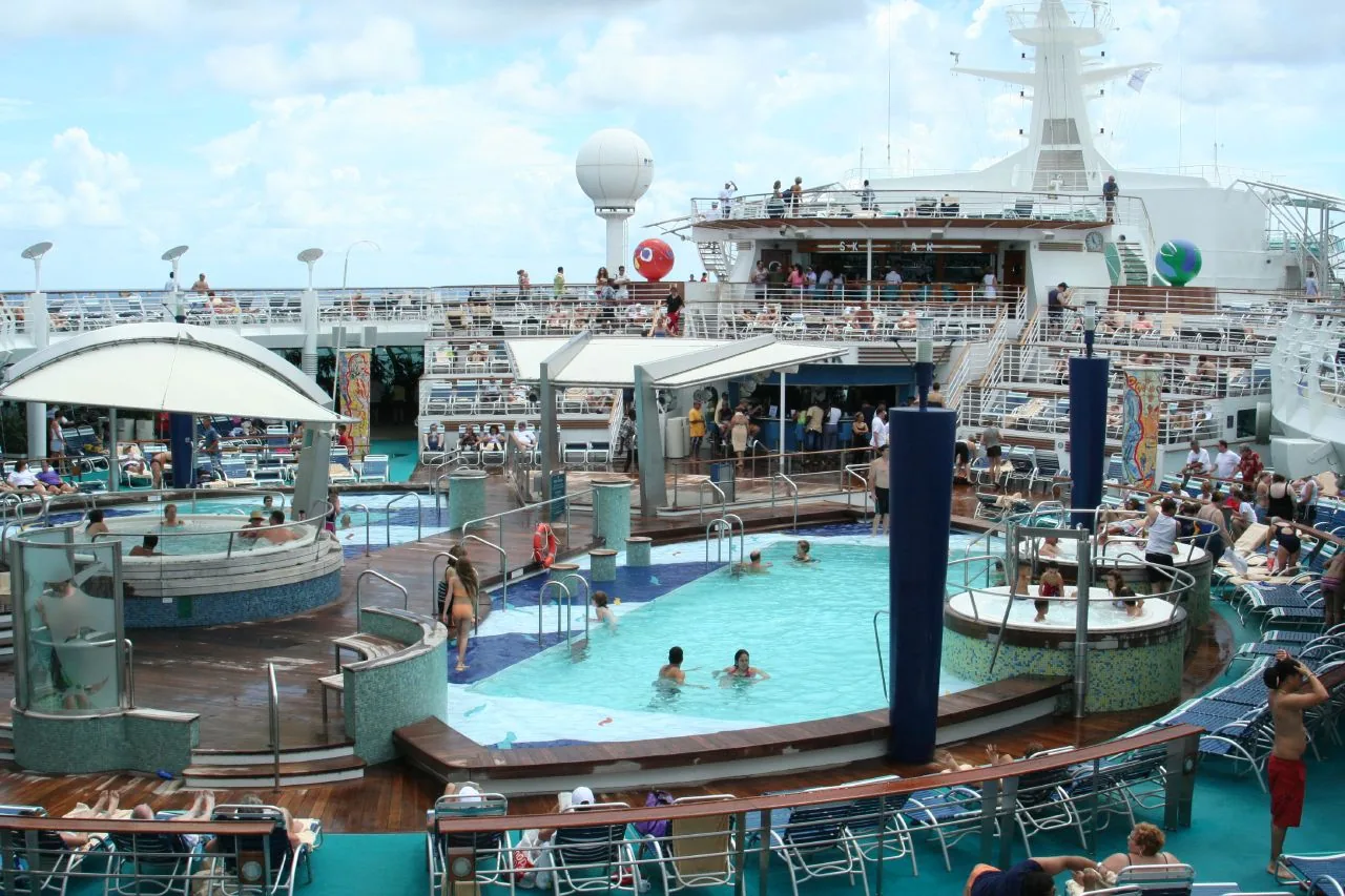 Busy cruise ship lido deck with multiple outdoor swimming pools, circular whirlpool spas, and rows of sunloungers filled with passengers on a sea day, with the ship's upper decks and bridge visible in the background