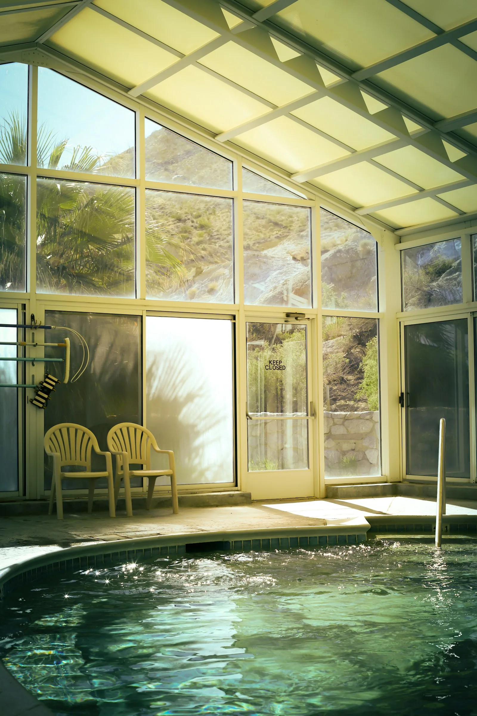 An enclosed indoor spa hydrotherapy pool with a glass and steel ceiling structure, green-tinted warm water with visible jet activity and a metal handrail entry step, representing the Oasis Spa thermal suite pool on P&O Britannia Deck 5