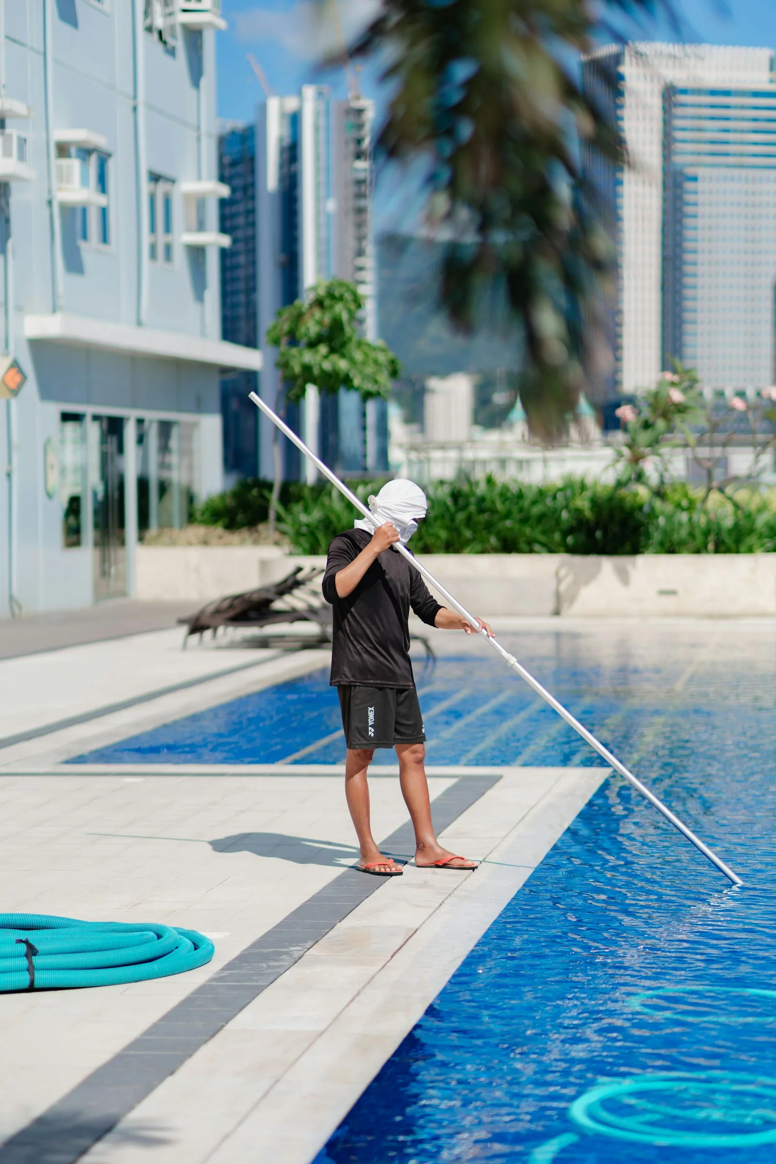 Pool maintenance technician standing poolside with a long-handled cleaning pole beside a hotel rooftop pool, representing pH minus application and chemical dosing technique