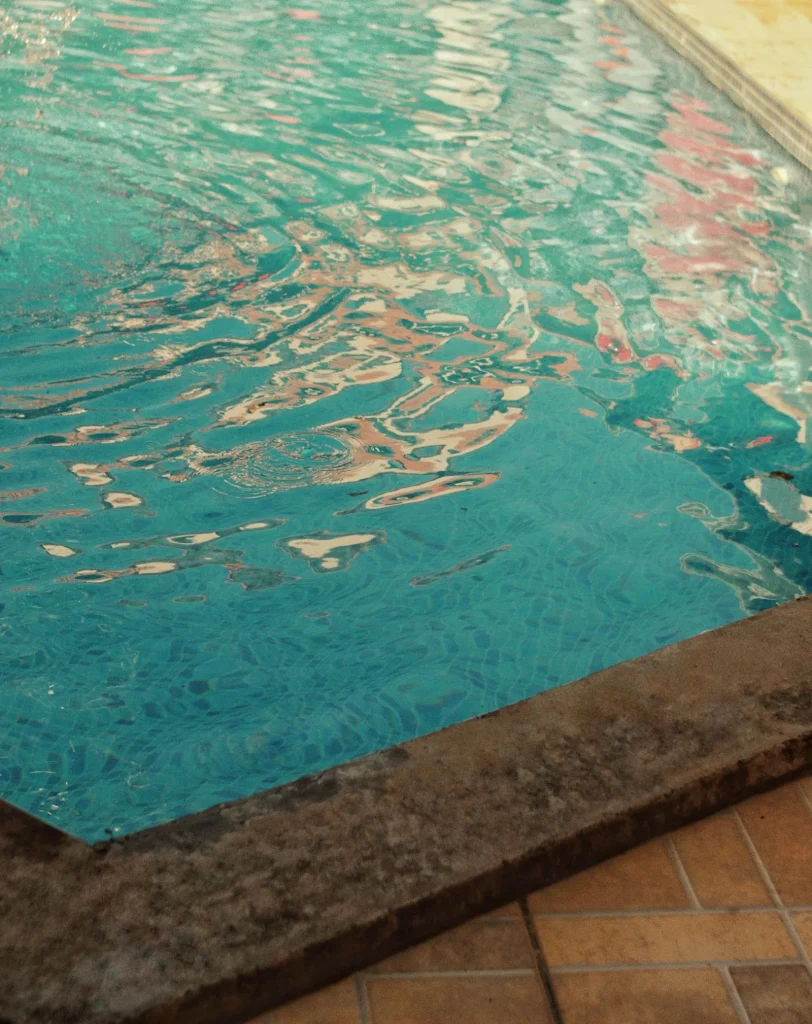 Close-up of clear turquoise swimming pool water reflecting light, with a stone pool coping edge and terracotta tile surround visible