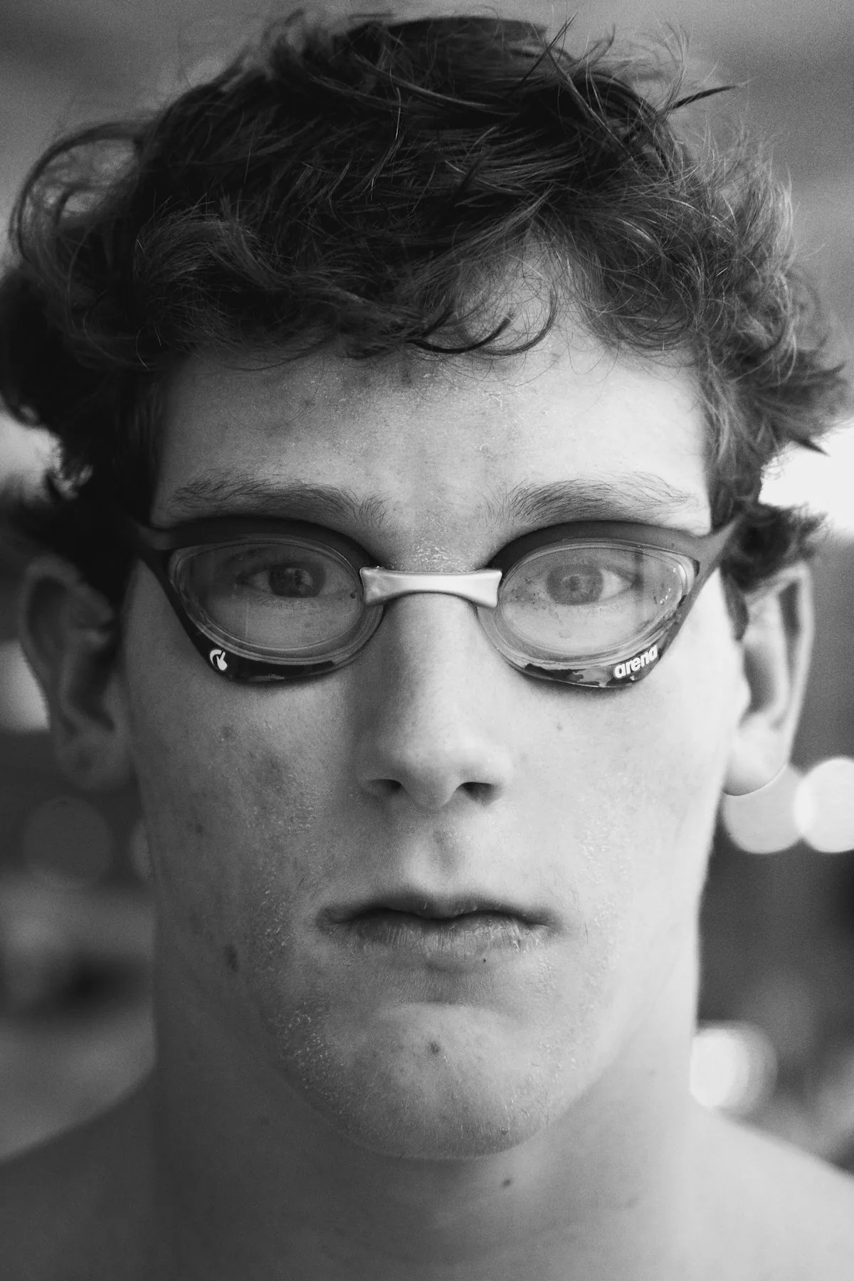 Close-up black and white portrait of a young male competitive swimmer wearing Arena racing goggles, representing the group most at risk from chlorine side effects due to prolonged indoor pool exposure