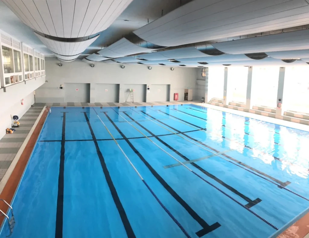 An empty indoor chlorinated swimming pool with lane markings and large HVAC ventilation ducts on the ceiling, representing the indoor pool environment where chloramine gas can accumulate above the water surface
