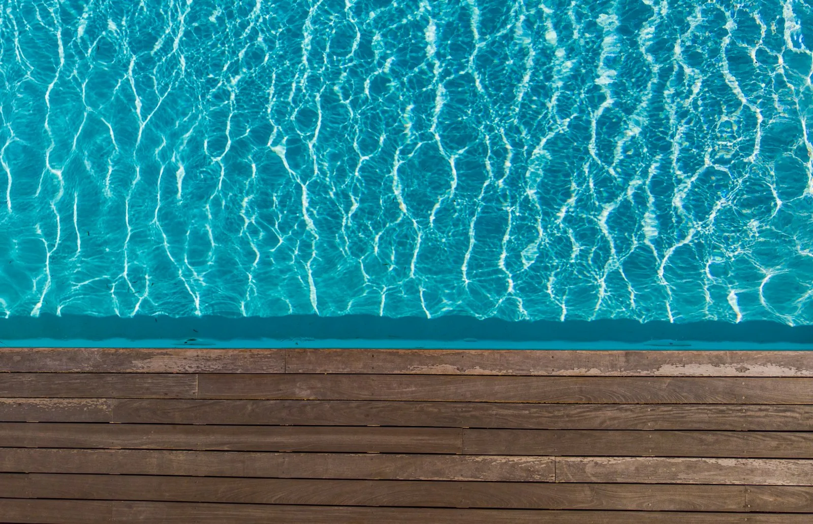 Close-up aerial view of clear turquoise pool water with light ripple patterns beside a wooden deck, representing a heated outdoor pool installation