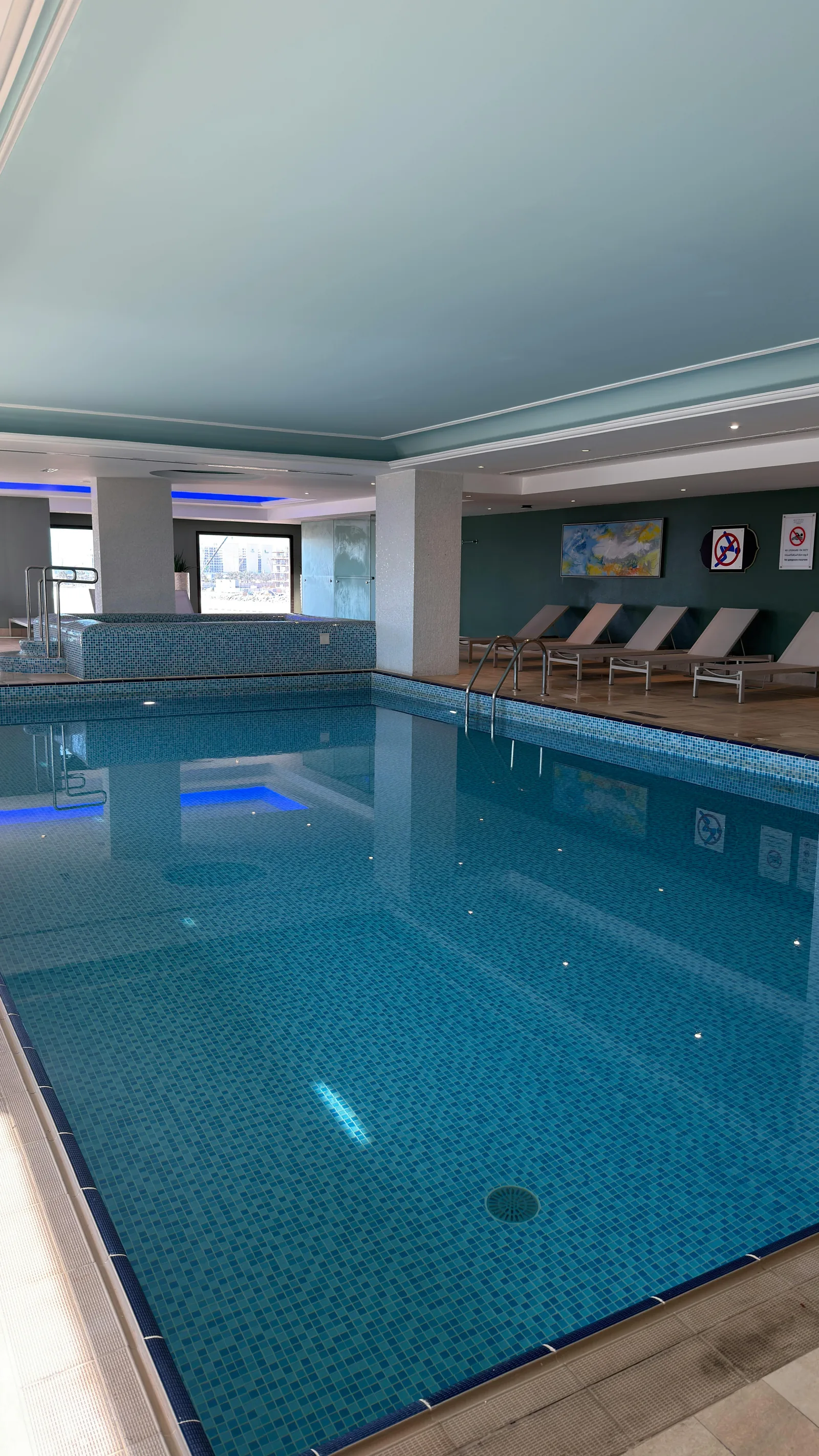 A modern indoor swimming pool with blue mosaic tiles, blue lighting and poolside loungers