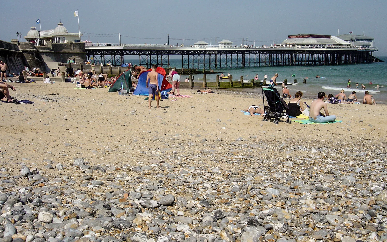 Cromer beach on a sunny summer day with the Victorian Pier and Pavilion Theatre visible in the background, people sitting on the mixed sand and shingle beach, and swimmers visible in the North Sea, representing sea swimming at Cromer on the Norfolk coast