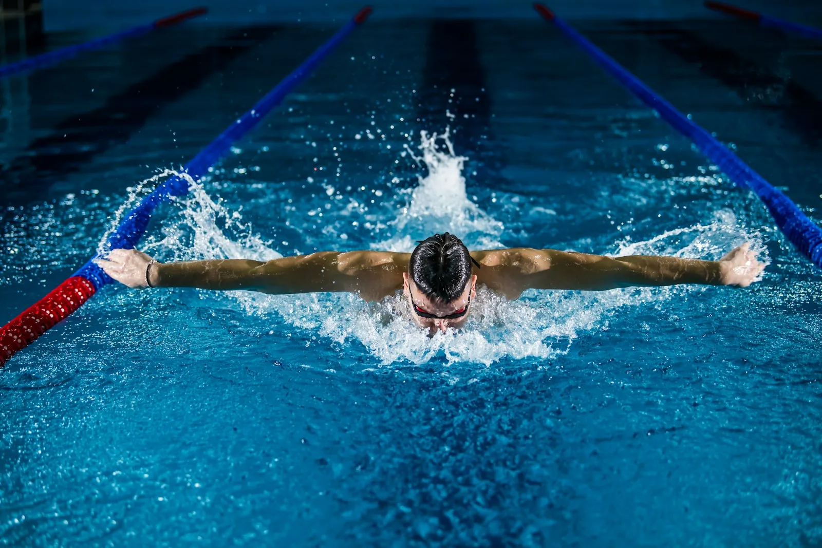Overhead view of a swimmer performing butterfly stroke in an indoor pool lane with red and blue lane ropes on either side, representing lane swimming at a public leisure centre such as Victory Swim and Fitness Centre near Cromer in Norfolk