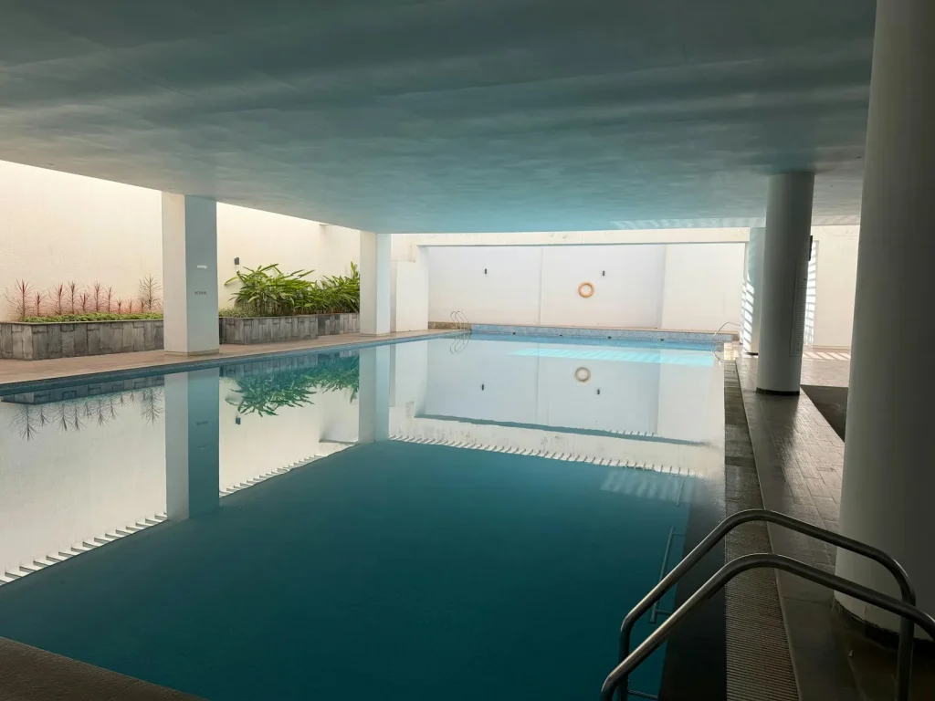 An indoor public swimming pool with still turquoise water, white walls, a lifering mounted on the wall, and a stainless steel handrail entry, representing the type of indoor 25-metre pool available at The Reef Leisure Centre in Sheringham near Cromer