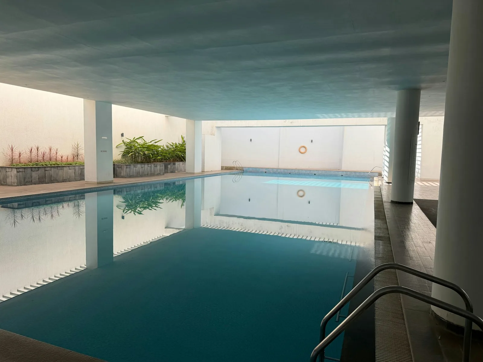 An indoor public swimming pool with still turquoise water, white walls, a lifering mounted on the wall, and a stainless steel handrail entry, representing the type of indoor 25-metre pool available at The Reef Leisure Centre in Sheringham near Cromer