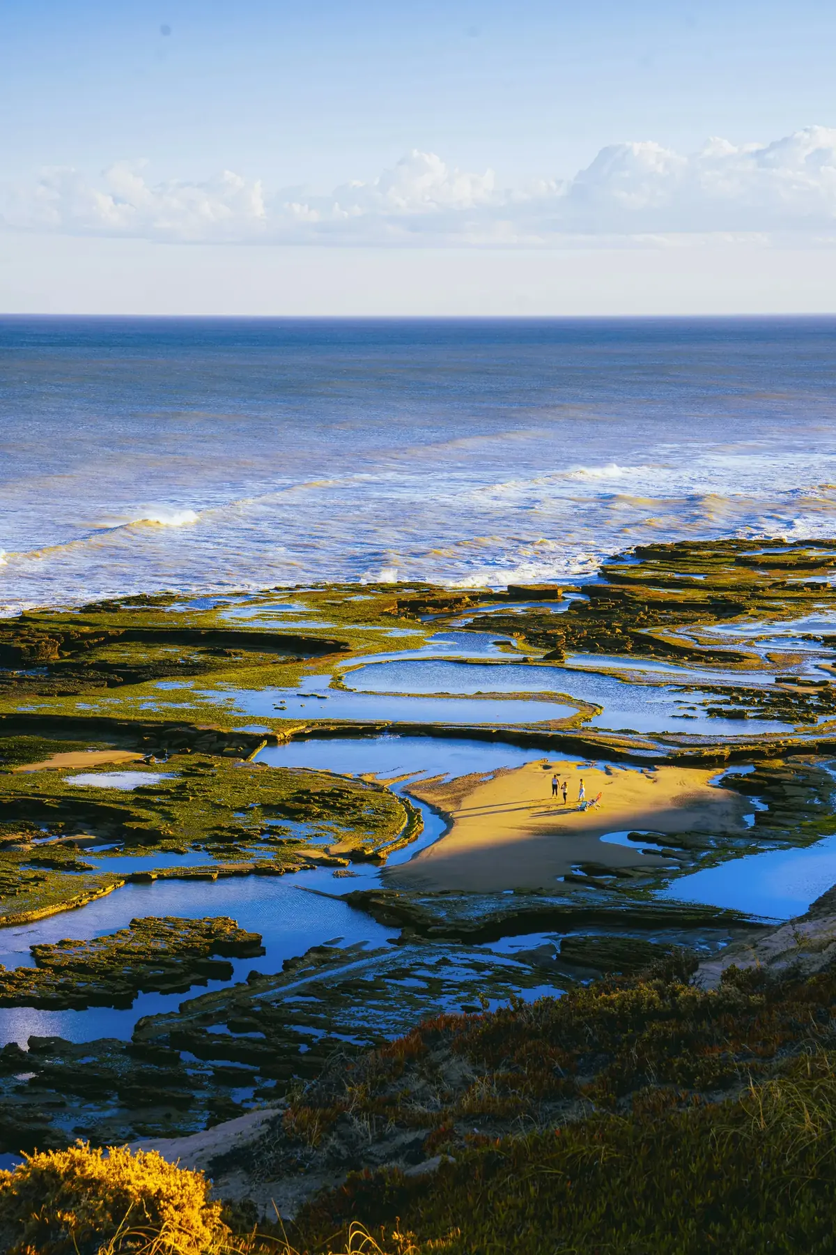 Aerial view of a rocky coastline with multiple natural tide pools at low tide, people walking among rock formations with ocean waves in background