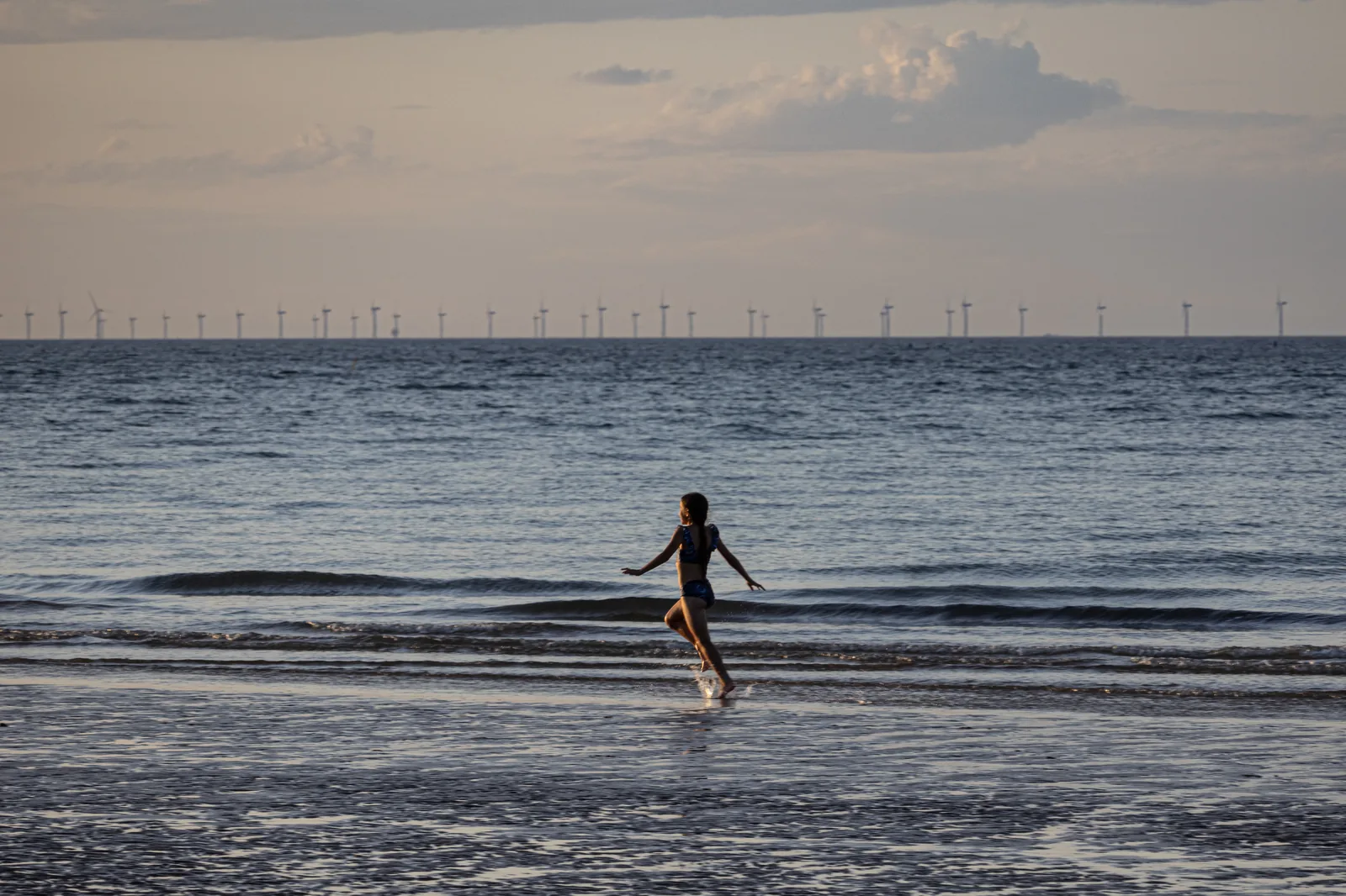 A person wading at West Runton beach with offshore wind turbines visible on the horizon, representing the North Norfolk coastal experience
